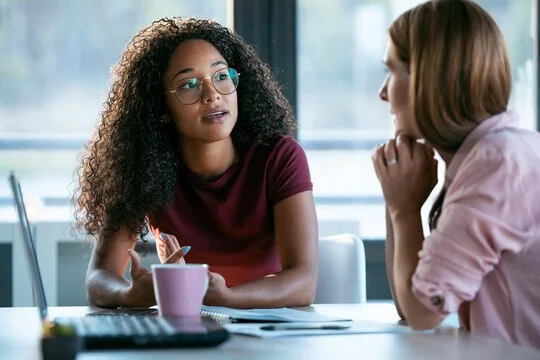 Two women having a conversation at a table in a bright room with large windows, one with curly hair and glasses, the other with straight hair.