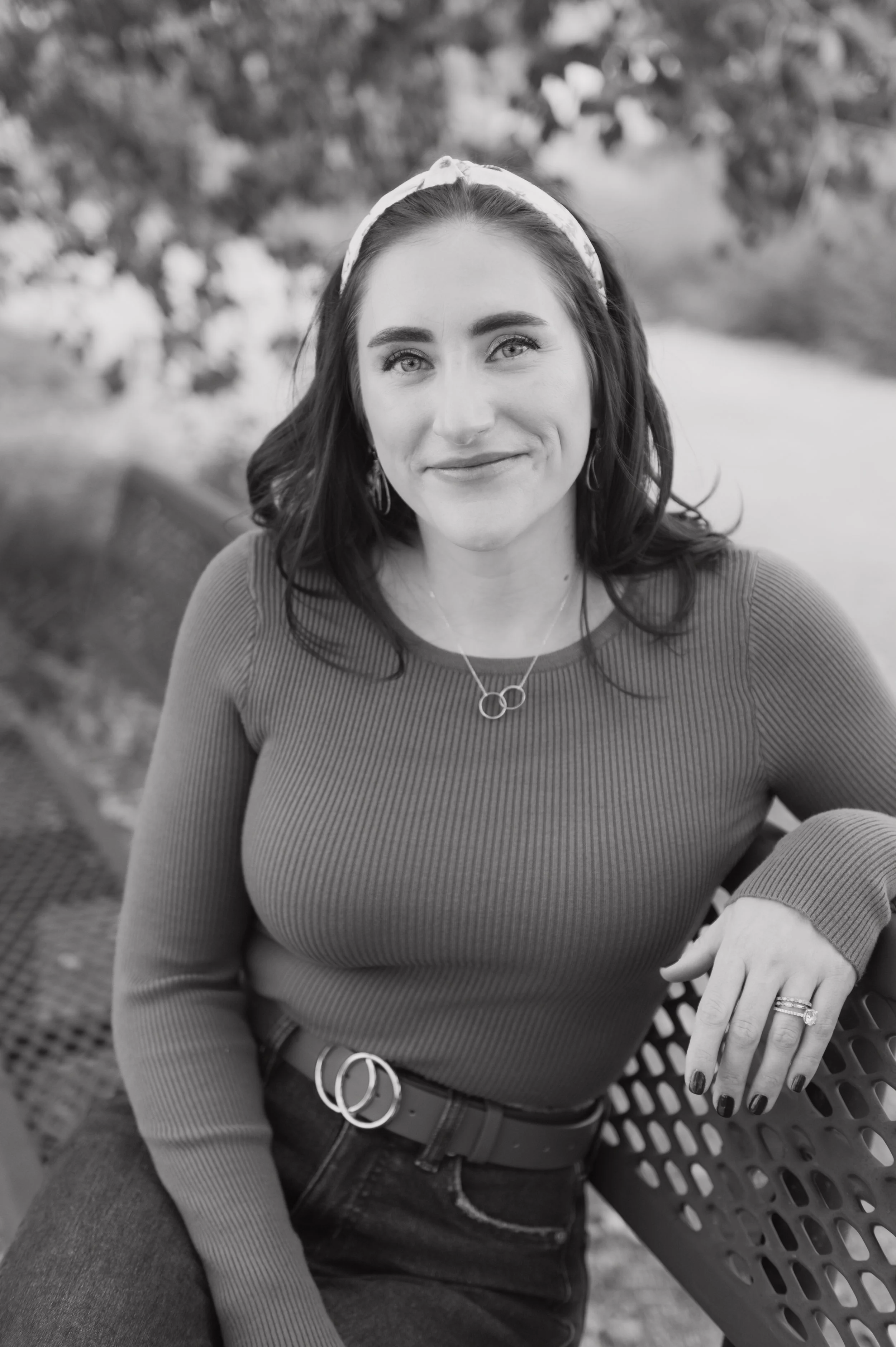 A woman with dark hair and light eyes, wearing a headband, smiling while sitting on a park bench. She has a ring, a necklace, and earrings, and is wearing a ribbed long-sleeve shirt and dark jeans.