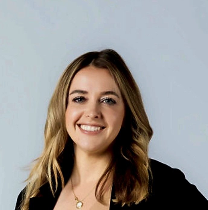 A woman with shoulder-length blonde hair smiling and wearing a black blazer and a gold necklace with a pendant, against a light gray background.