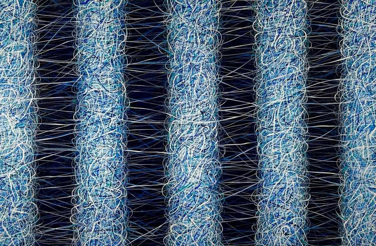 Close-up of a cylindrical object made of tangled light blue and white threads or wires, arranged in vertical columns against a dark background.
