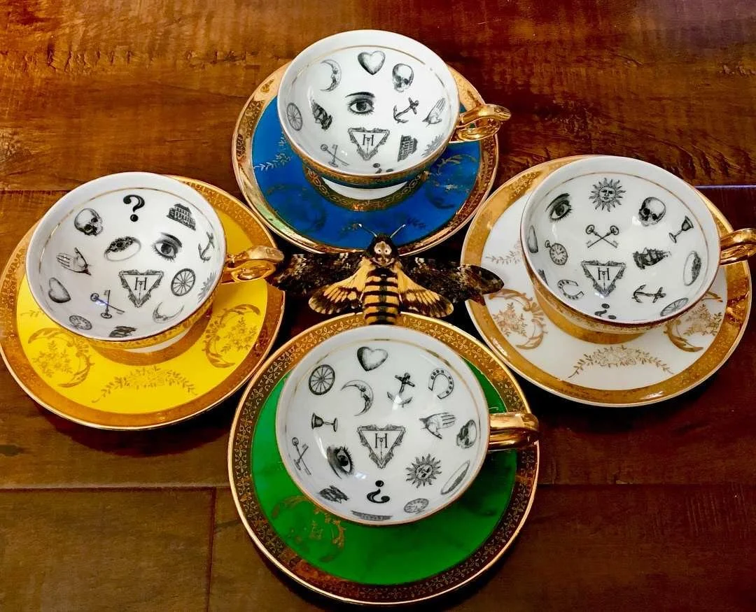 Four ornate teacups with matching saucers, decorated with various mystical symbols and illustrations, arranged in a circular pattern on a wooden surface, with a decorative bee or insect figurine in the center.