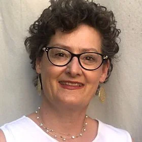A mature woman with curly blonde hair and glasses resting her face on her hand, wearing a black sheer blouse with pink floral patterns, earrings, and a smartwatch, sitting in front of a wood-paneled background.