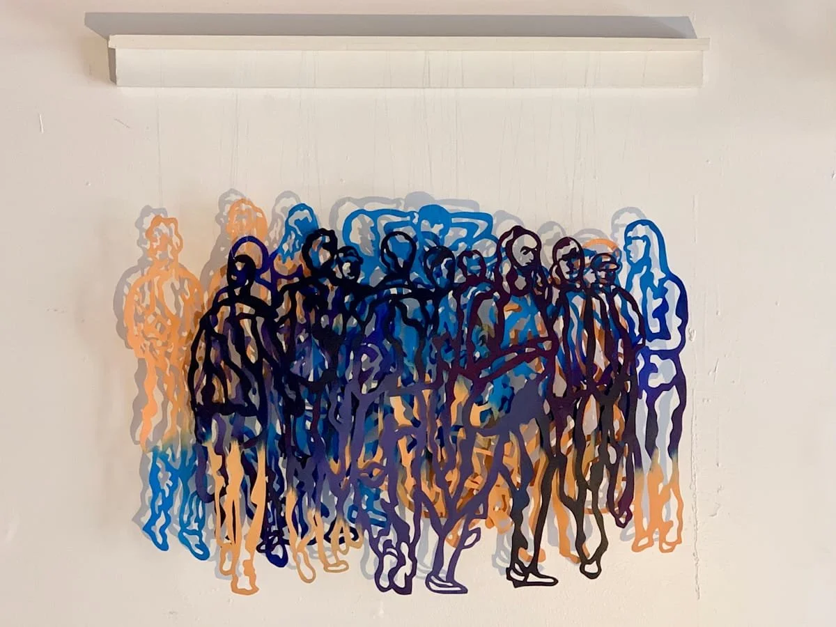 Colorful wall art depicting a group of people outlined in black, with some figures in orange, blue, and purple, on a white wall.