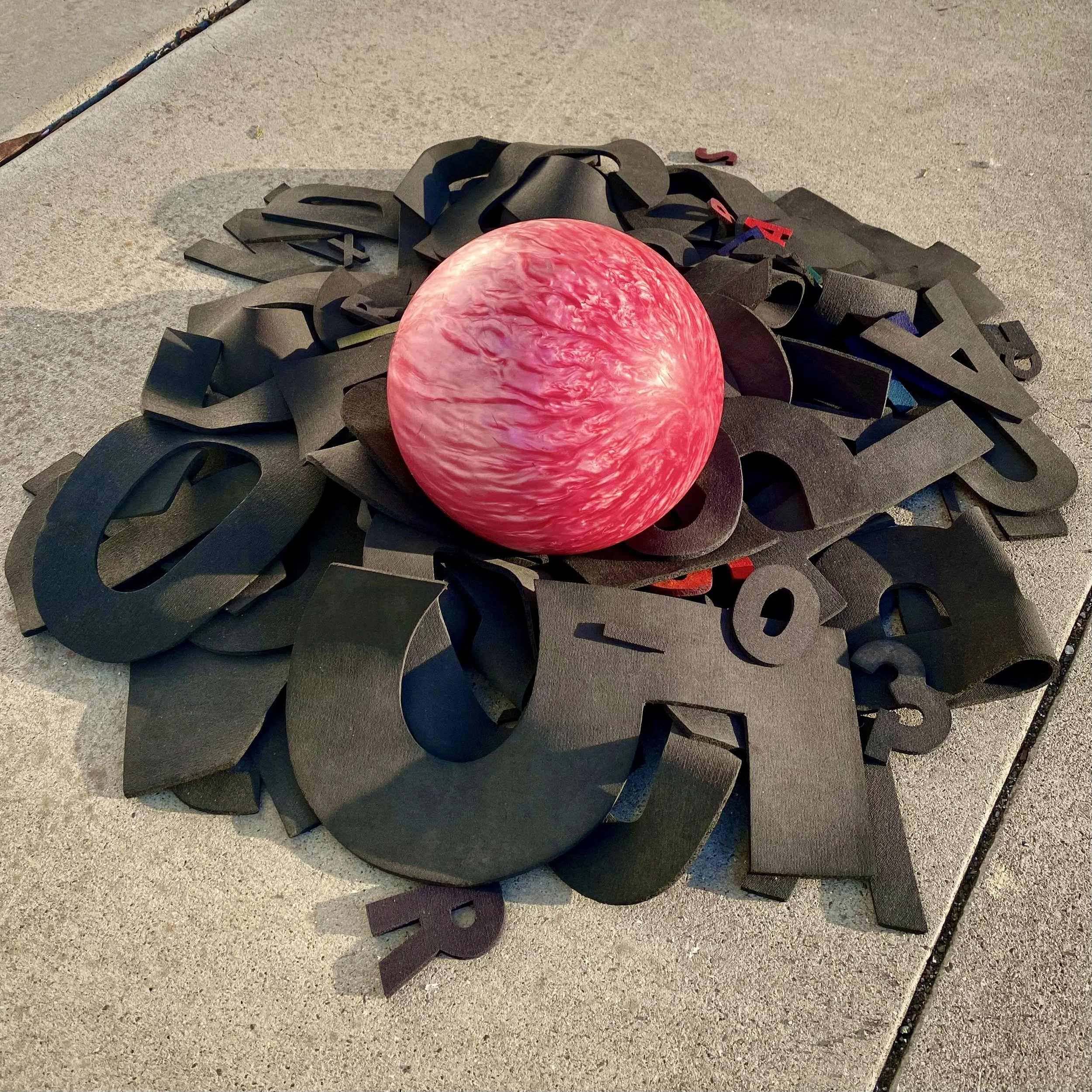A large, red and white marbled sphere sculpture placed on a pile of black metal cutouts of letters and numbers on a concrete surface.