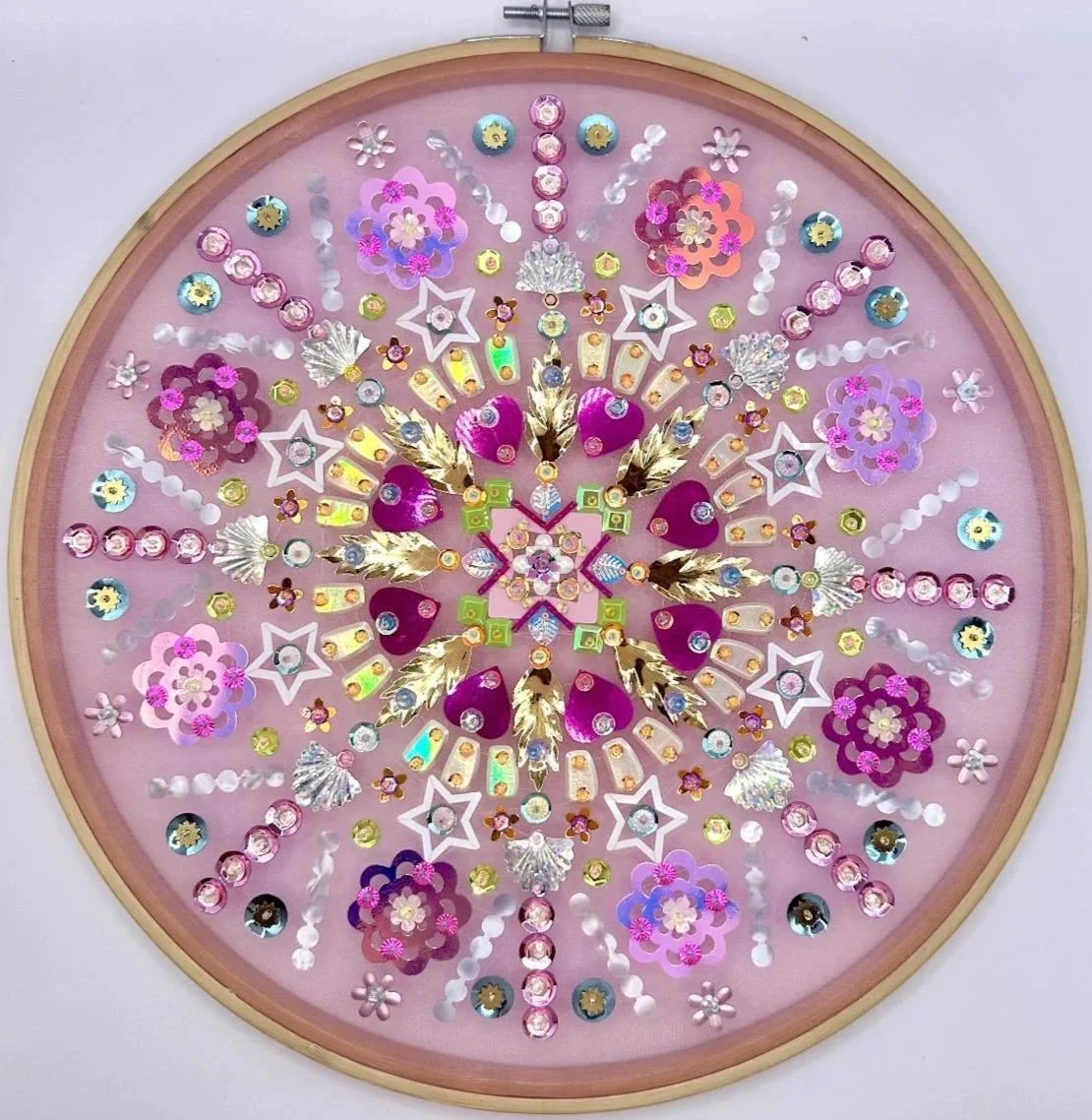 Circular decorative collage with pink background, featuring various rhinestones, sequins, and small decorative elements arranged in a symmetrical pattern.