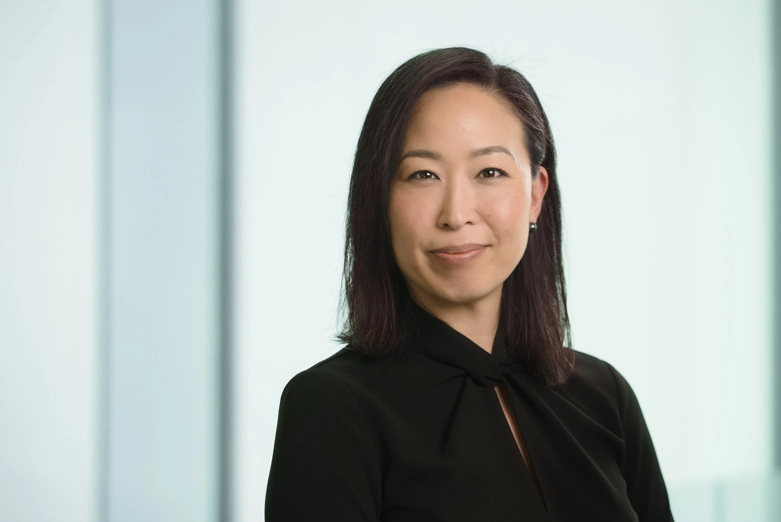 A professional woman with shoulder-length dark hair, wearing a black top with a small bow, standing in front of a light-colored background with vertical lines.