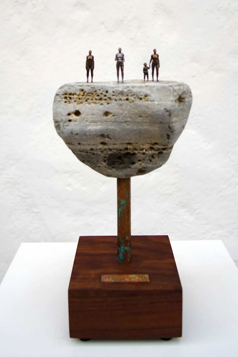 Miniature sculpture of a large rock with tiny people figurines standing on top, mounted on a wooden base.