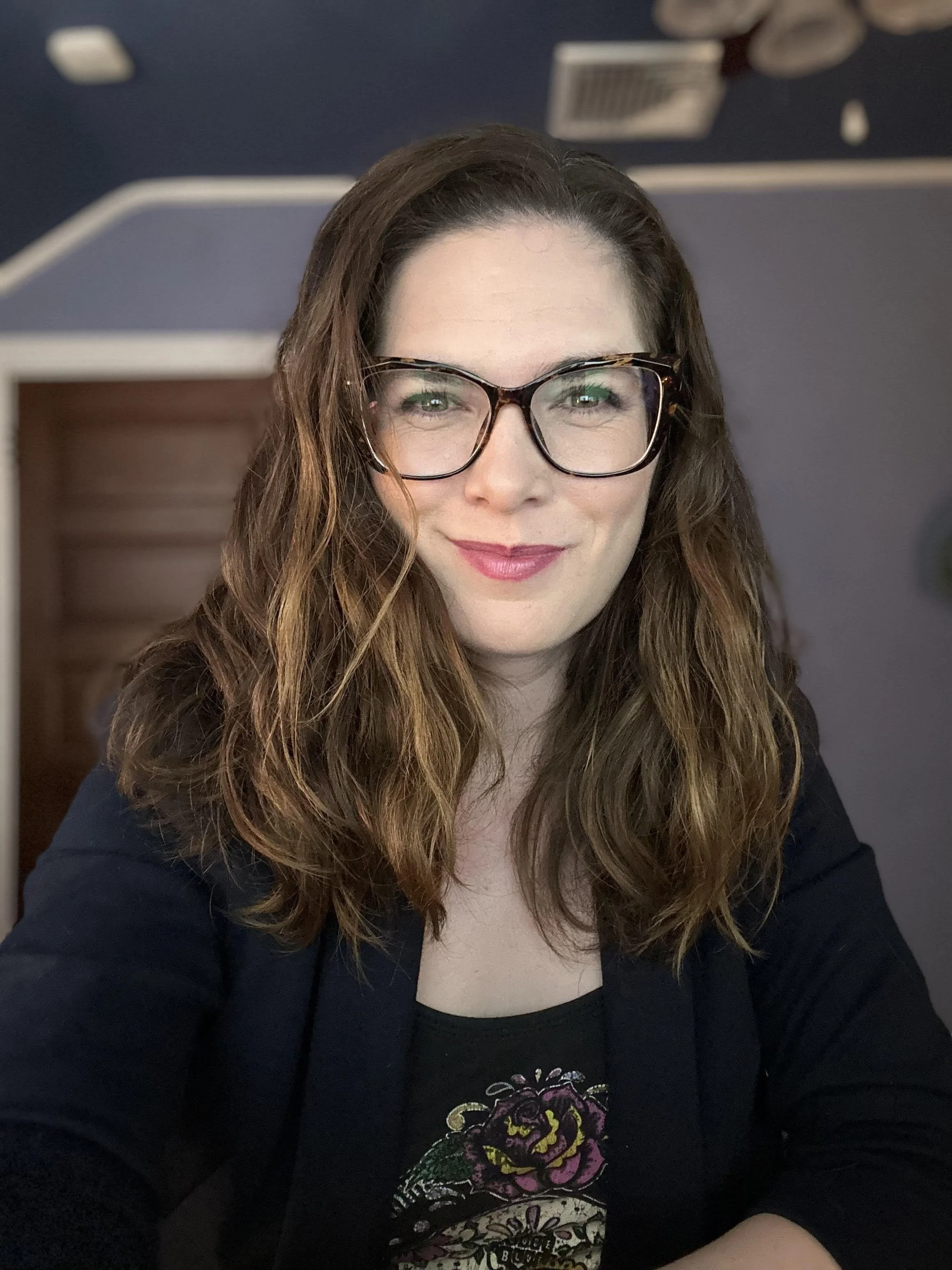 A woman with long wavy brown hair, wearing black glasses and a red patterned top, smiling in a room with warm lighting.
