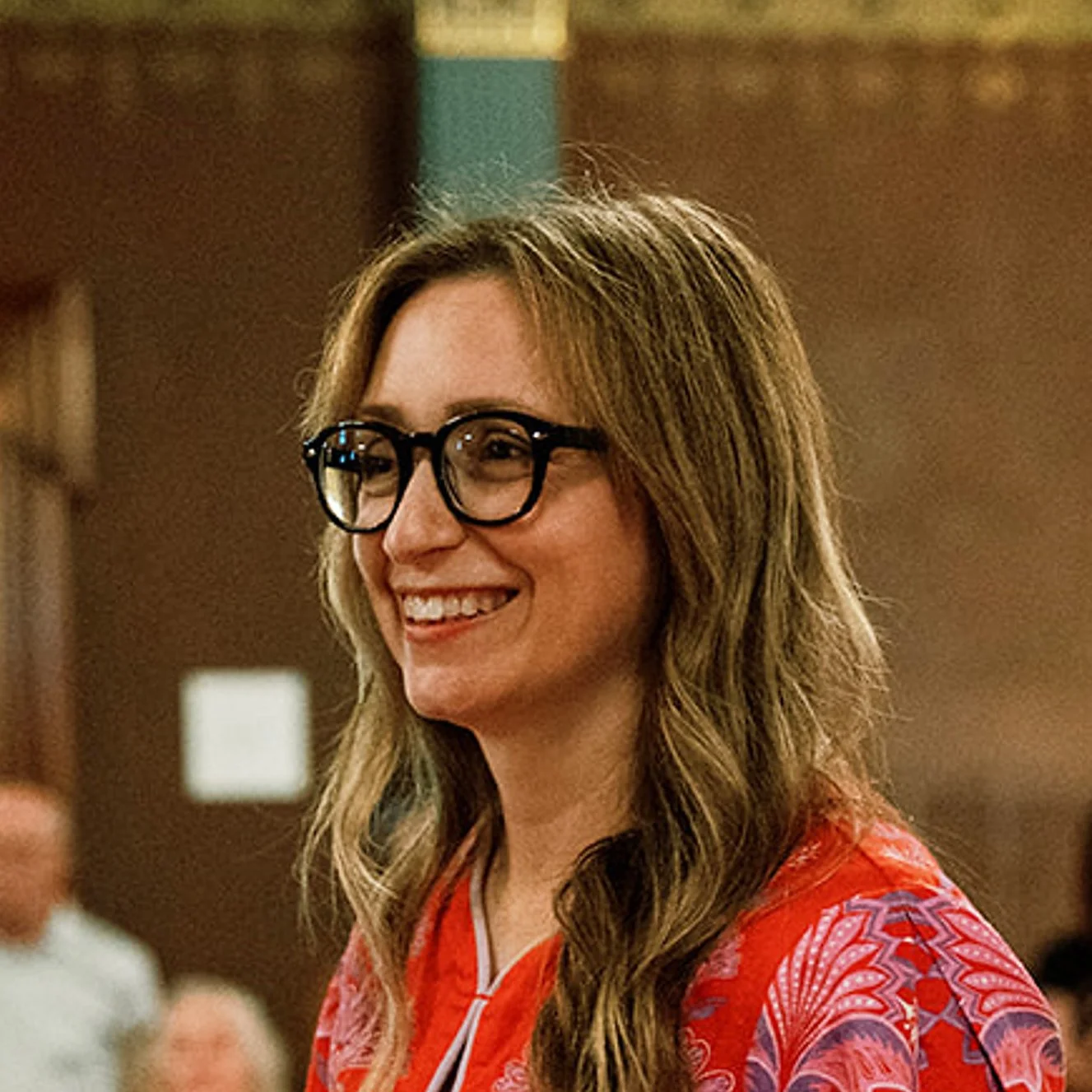 A woman with long wavy brown hair, wearing black glasses and a red patterned top, smiling in a room with warm lighting.