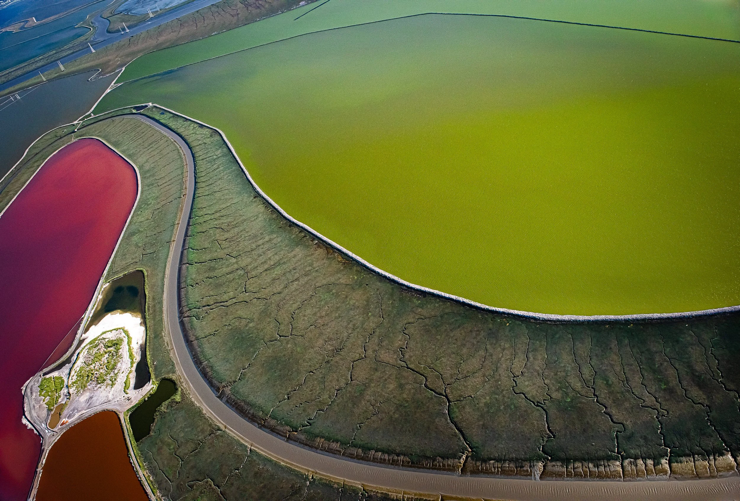 Aerial view of colorful salt evaporation ponds with vibrant green, red, and brown hues, divided by paths and barriers.