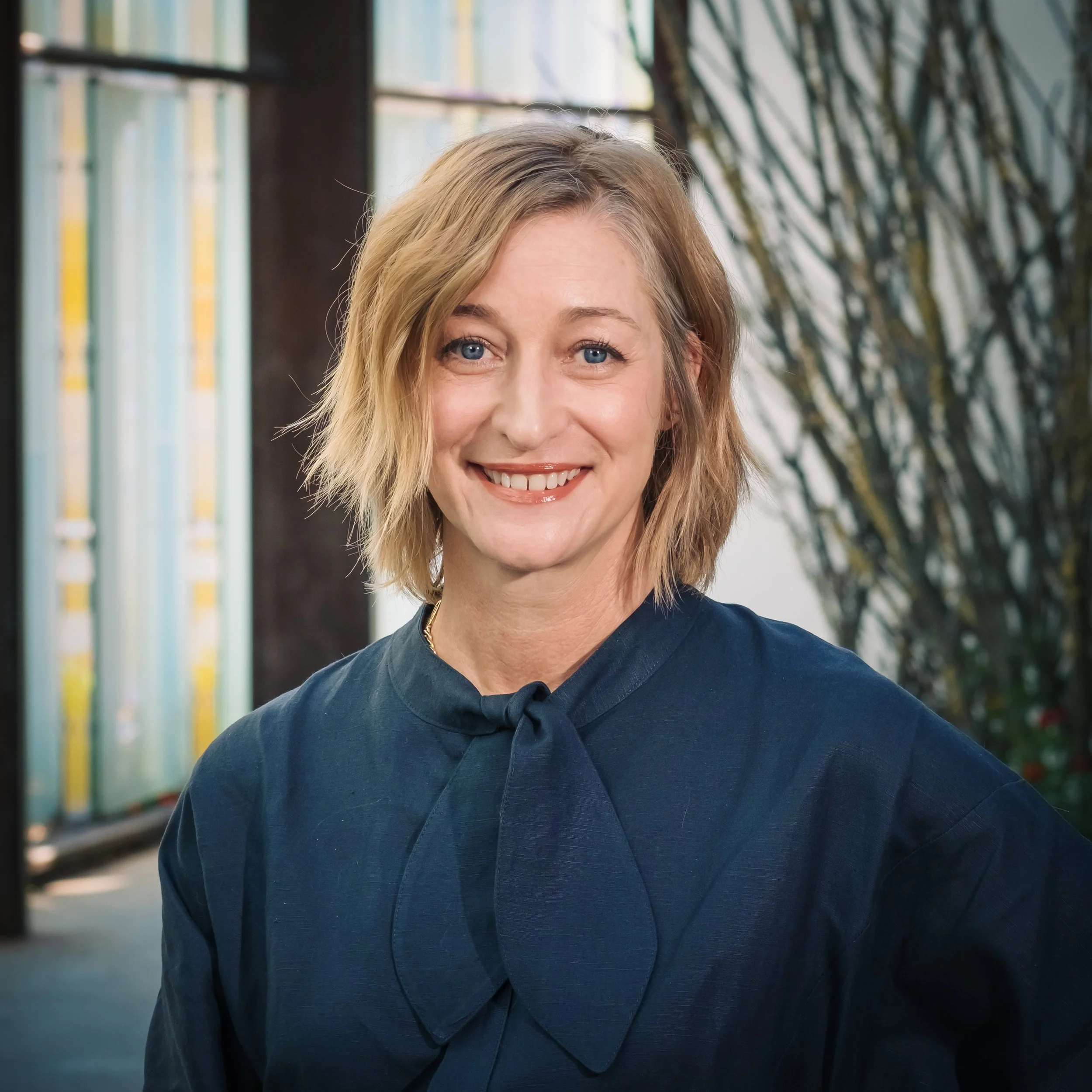 A woman with short blonde hair, blue eyes, and a friendly smile, wearing a navy blue blouse with a bow tie, standing indoors with large windows and greenery in the background.