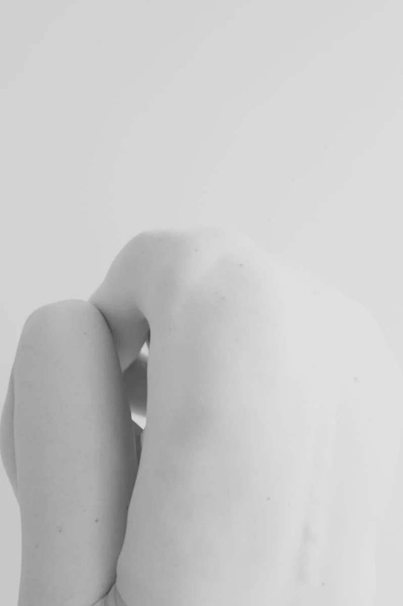 Close-up of a person’s back and shoulder, with their arm bent and hand resting on their shoulder. The image is in black and white.