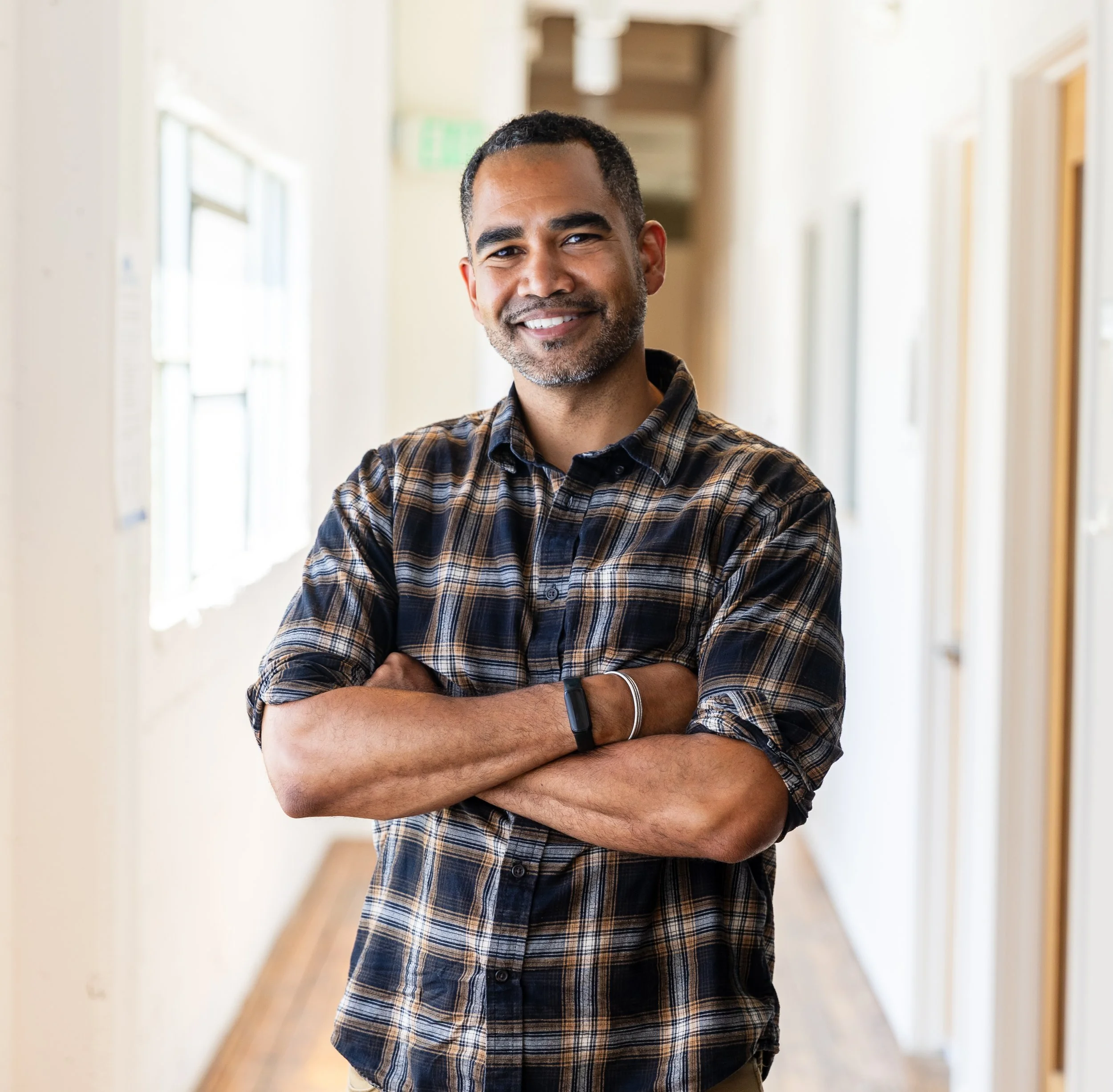 A smiling man with short dark hair, beard, and wearing a plaid shirt, stands with arms crossed in a bright hallway with windows and doors.
