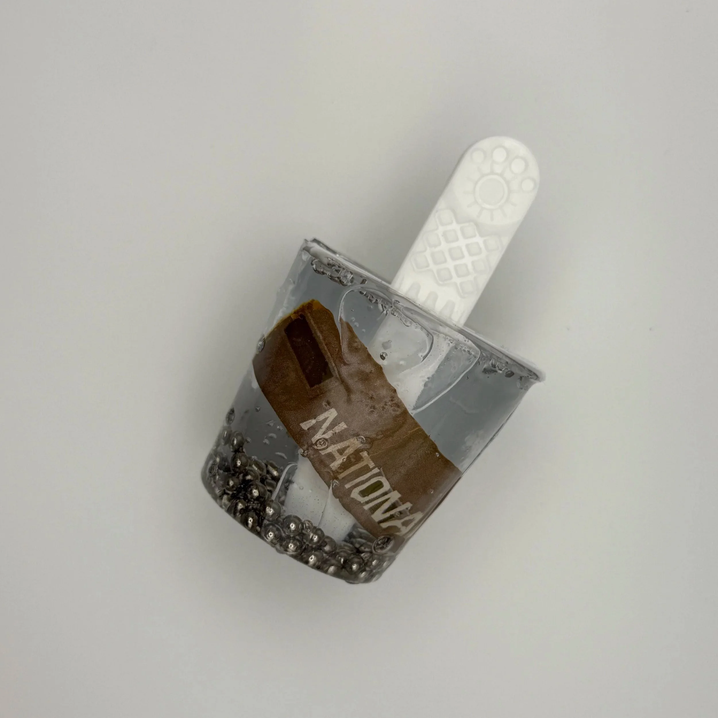 A clear glass of water with a brown paper label and metal balls at the bottom, a white paddle with a textured handle sticking out of the glass.