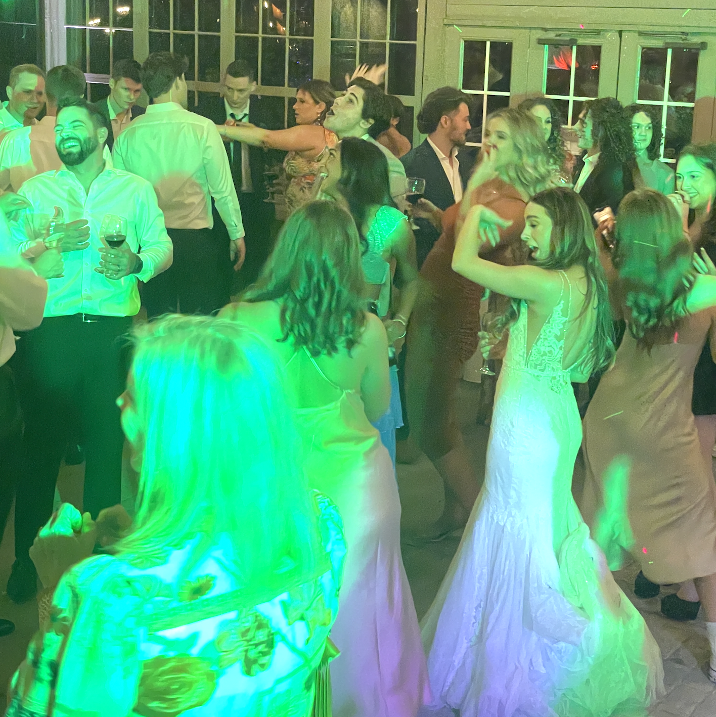 People dancing and socializing at a wedding celebration, with a woman in a white dress and others in formal attire, under green and colorful lighting.
