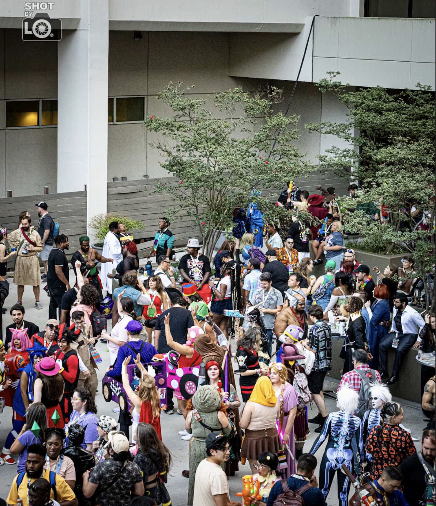Large group of people in colorful costumes, some with props, gathered outdoors near trees and modern buildings at a convention or festival.