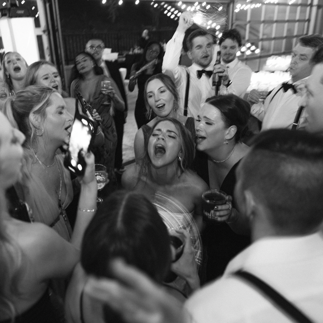 A group of people at a lively wedding, singing and dancing closely together, with some holding drinks, in a festive setting with string lights overhead.