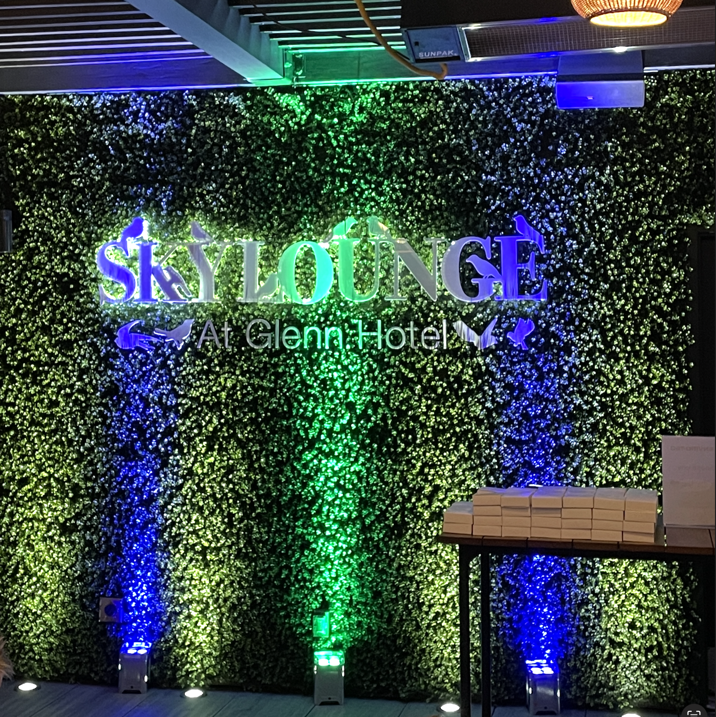 Neon sign reading 'Sky Lounge at Glenn Hotel' against a backdrop of lush green and white plants, illuminated by multicolored up-lighting.