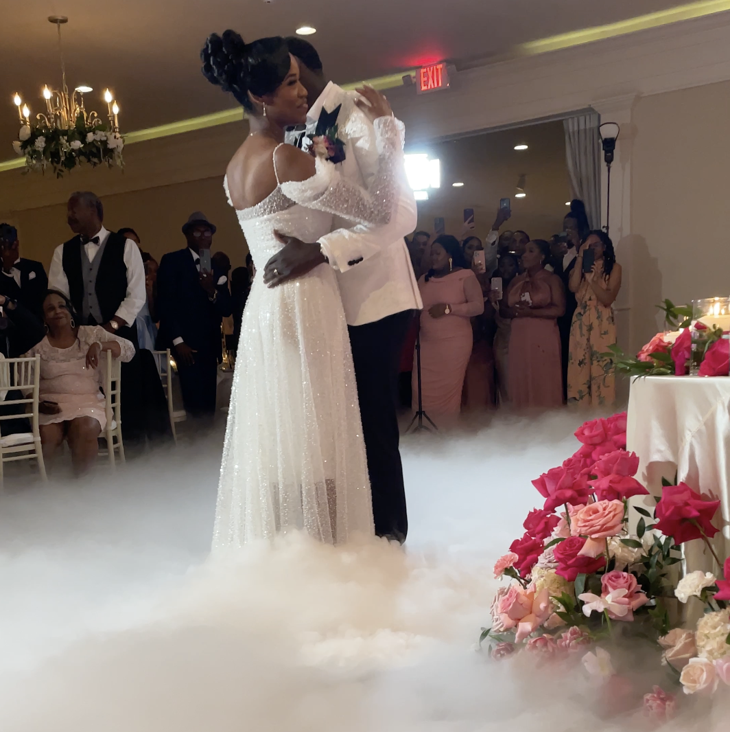 A bride and groom sharing a dance at their wedding reception, surrounded by guests, with pink flowers on a table to the right and fog on the floor.
