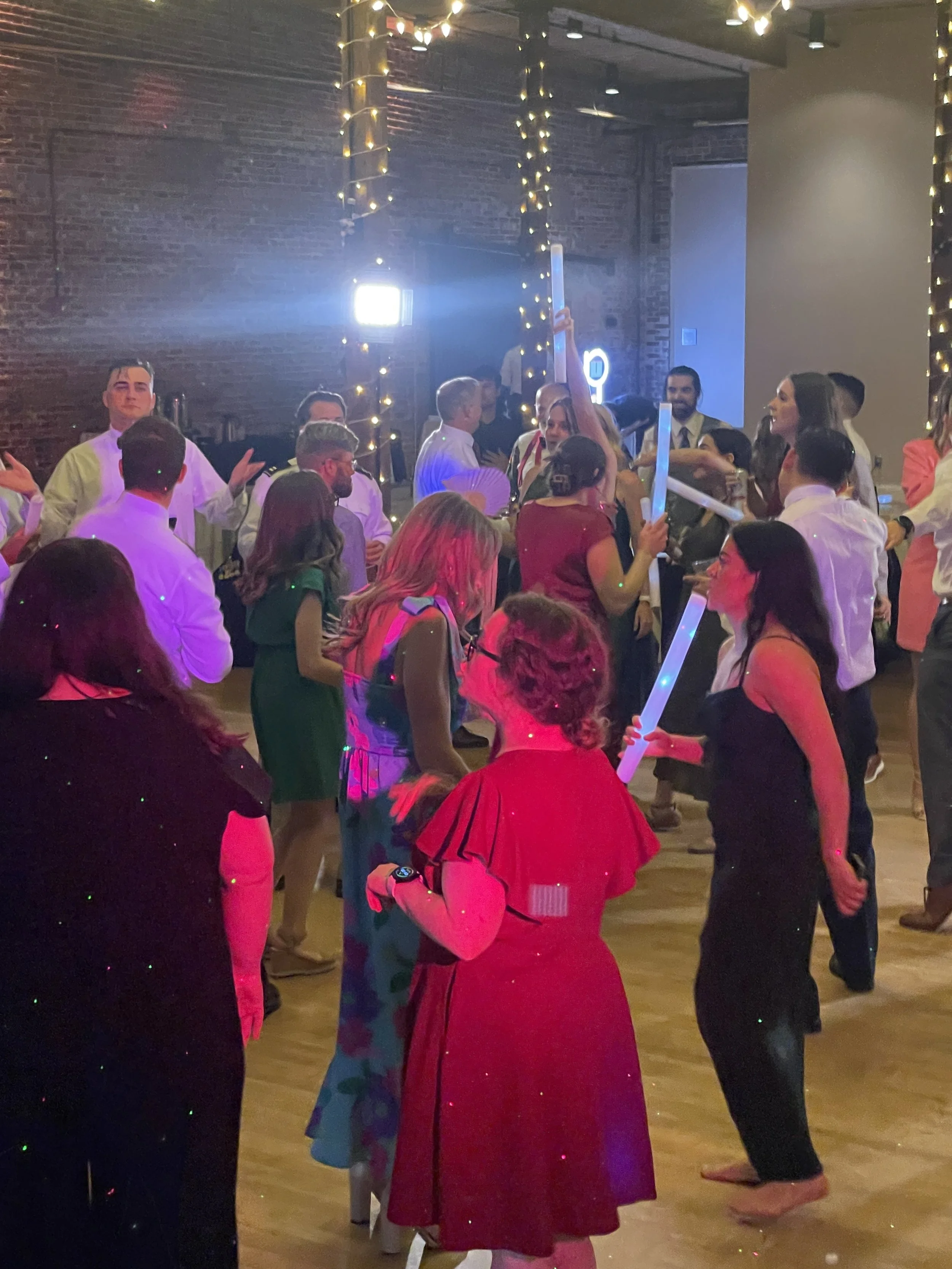 People dancing and celebrating at a party or wedding reception in a decorated venue with string lights and exposed brick walls.