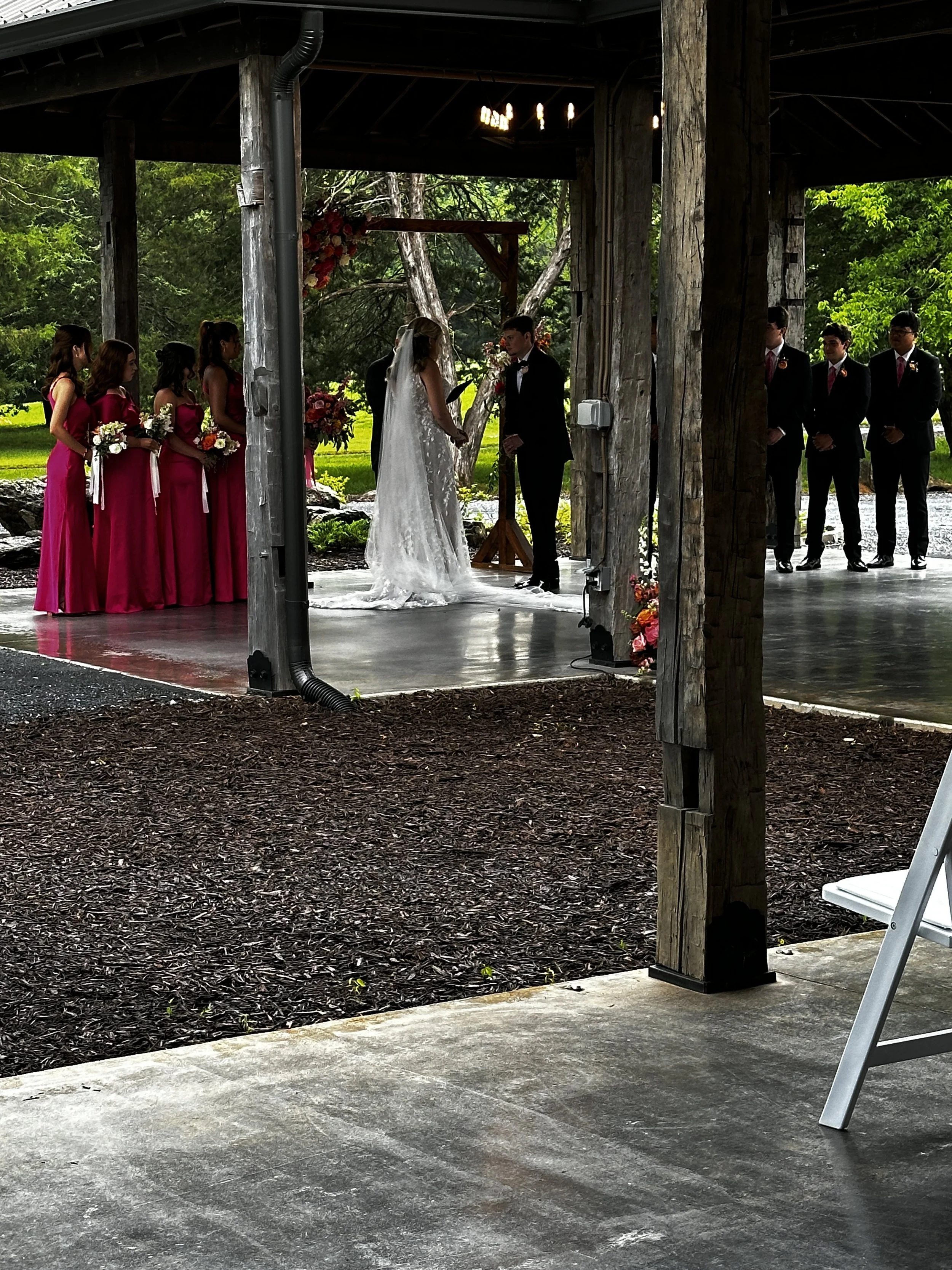 A wedding ceremony is taking place under a rustic wooden structure in a park. The bride and groom stand in front, holding hands, with the bride in a white dress and the groom in a black suit. Bridesmaids in pink dresses stand on the left, holding bouquets, and groomsmen in dark suits with pink ties stand on the right. Green trees and grass surround the ceremony area.