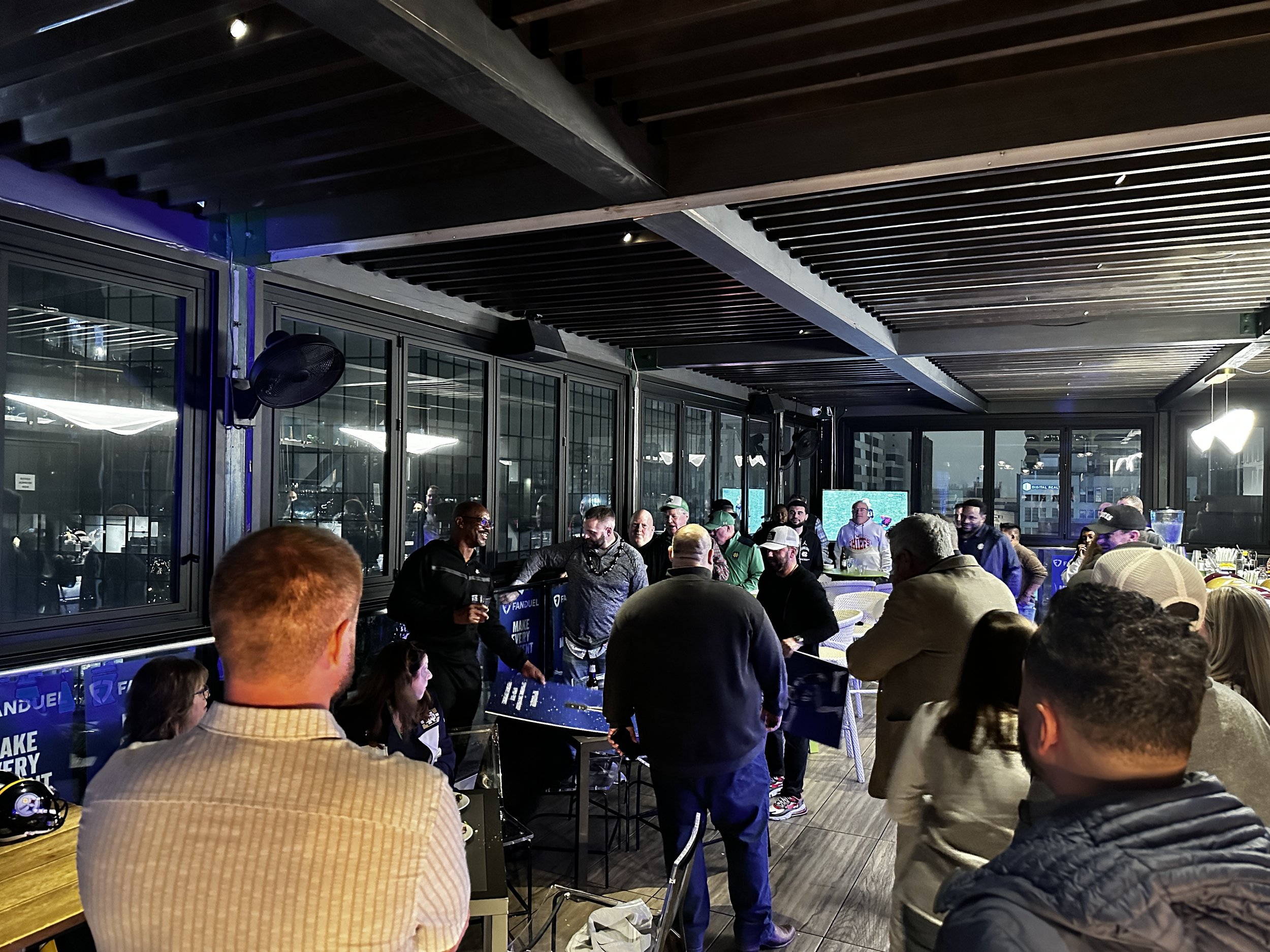 People gathered at a social event or party in an indoor space with large glass windows, some standing and talking while others watch a vendor or announcer at a table. The room has a modern design with a dark ceiling and bright lighting.