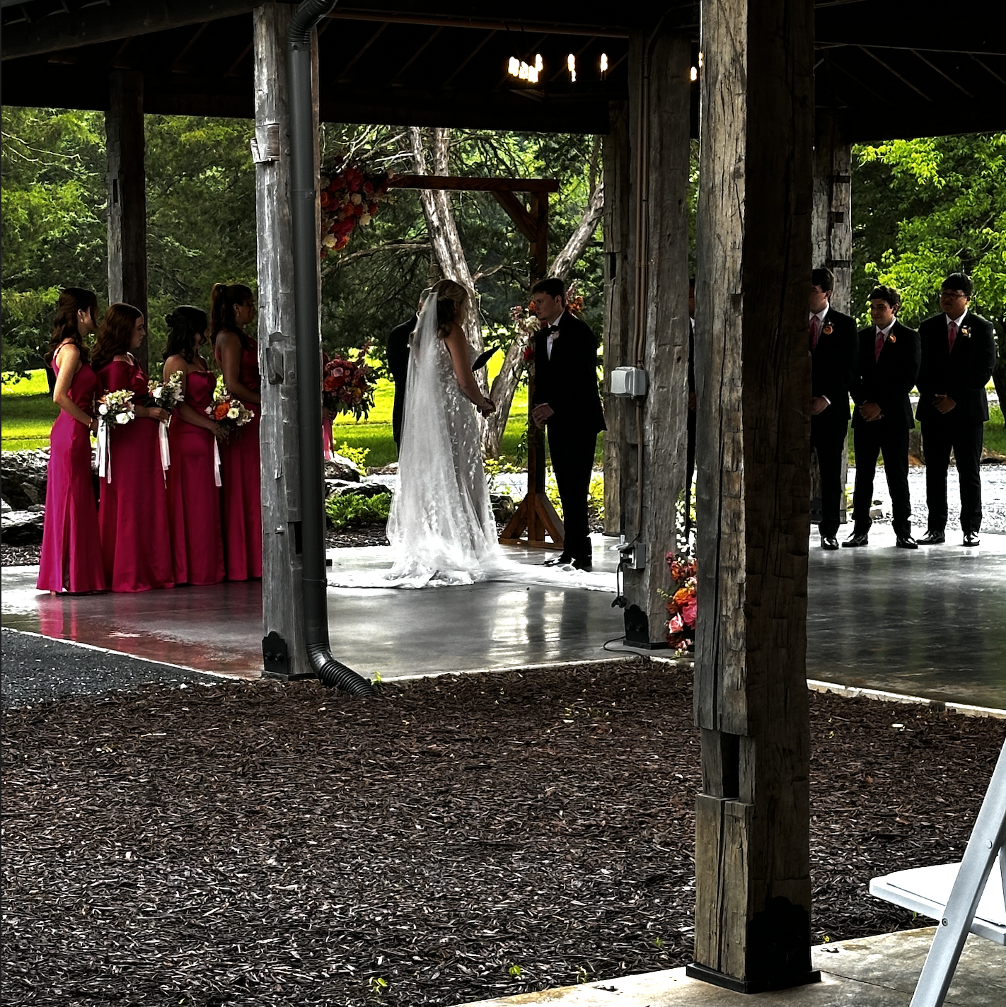 A wedding ceremony taking place outdoors under a wooden shelter, with a bride and groom holding hands at the altar, surrounded by bridesmaids in pink dresses and groomsmen in black suits, with greenery and trees in the background.