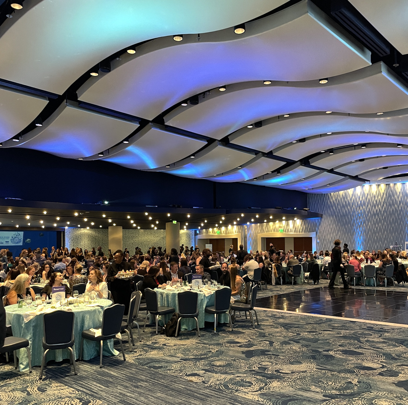 Large banquet hall filled with round tables and seated attendees at a formal event, with decorative ceiling and wall lighting.