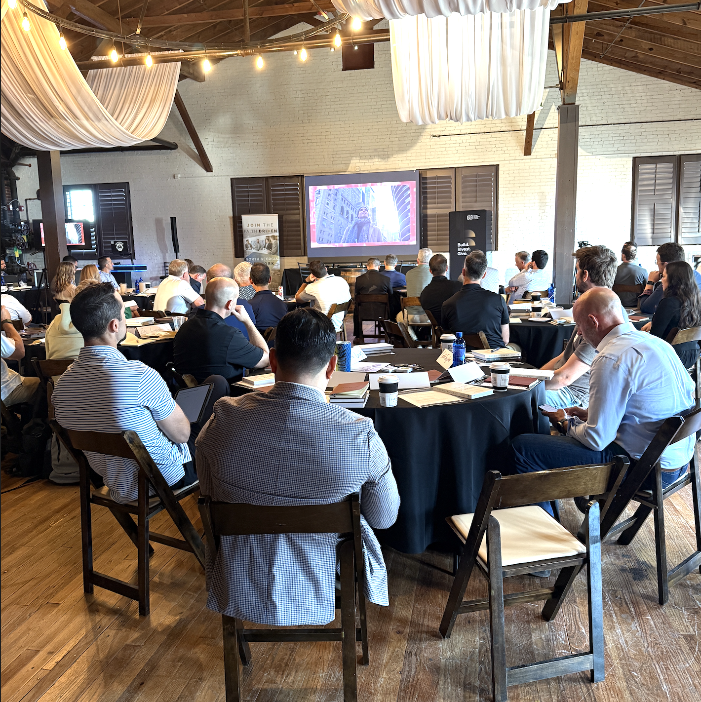 Group of people attending a conference or seminar in a large, rustic room with exposed brick walls and wooden ceiling beams, seated around round tables, watching a presentation on a large screen.