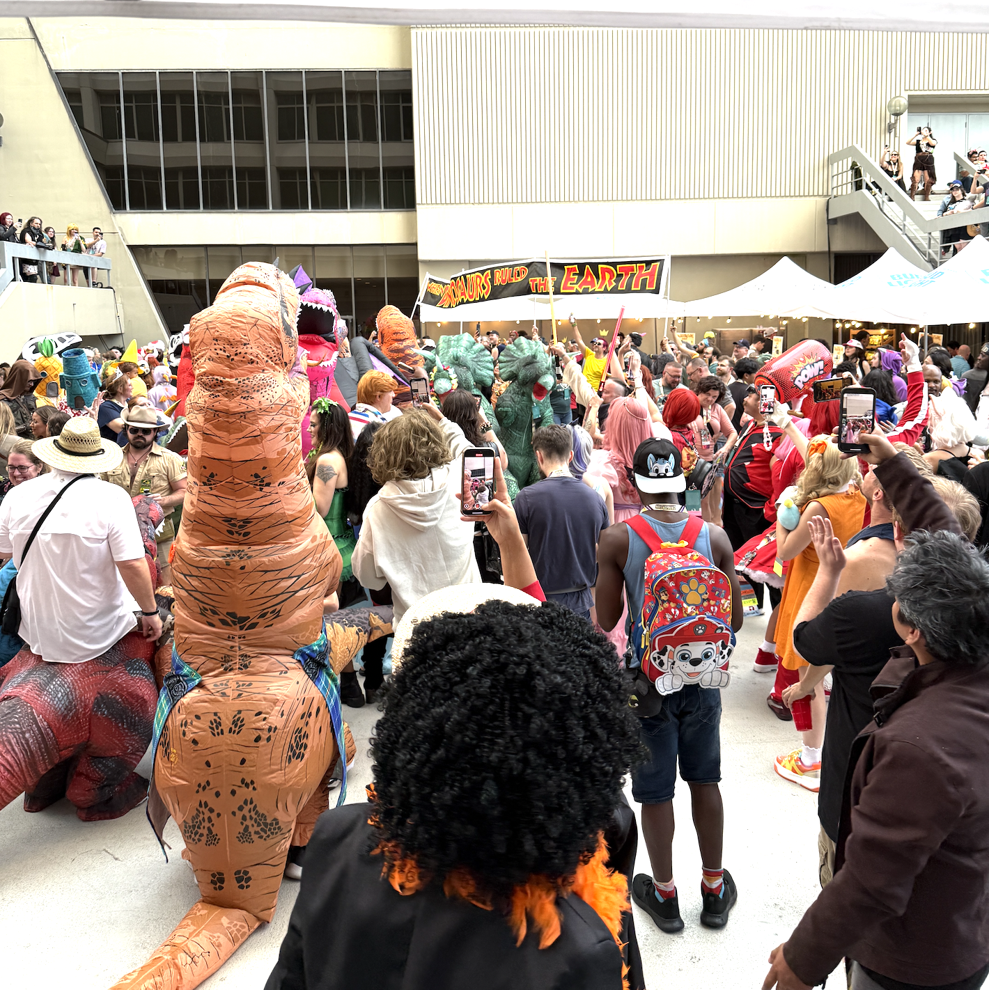 Crowd of people attending DragonCon in Atlanta dressed as dinosaurs and other characters with a banner reading "Dinosaurs Ruled the Earth" in the background.