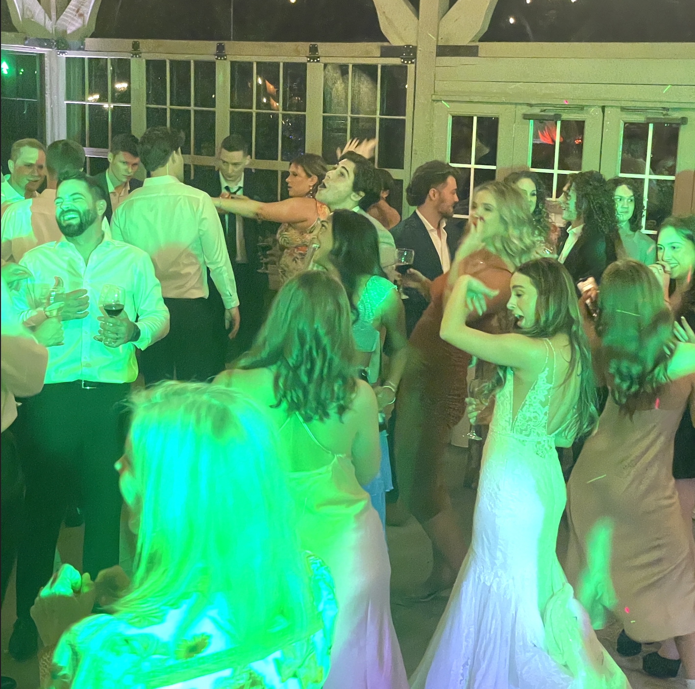 Group of people dancing and socializing at a wedding, some holding glasses of wine, with green lighting and a wooden interior.