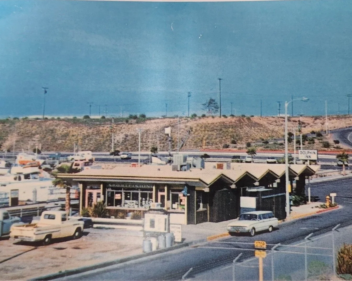 A vintage photo of a commercial area with a small market, vintage cars, and a highway in the background under a blue sky.