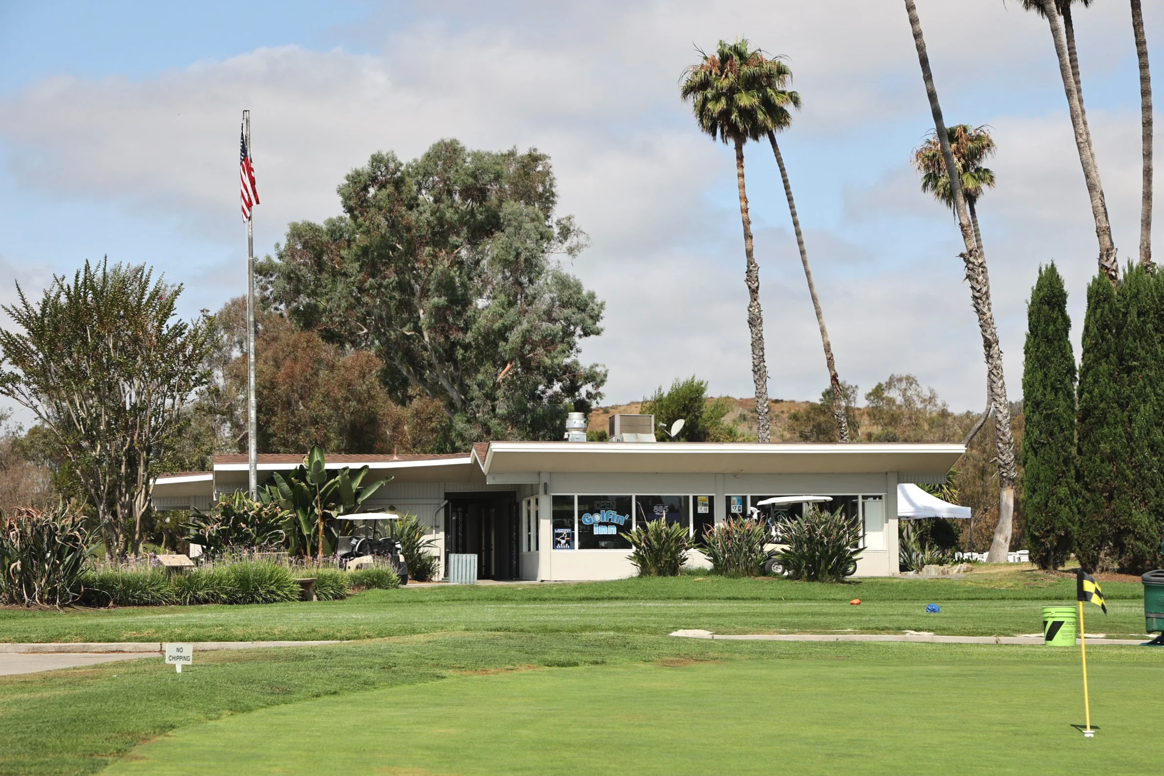 Oceanside Golf Course restaurant The Golfin' Inn with large front windows, surrounded by palm trees and greenery, with golf flags and a putting green in the foreground.