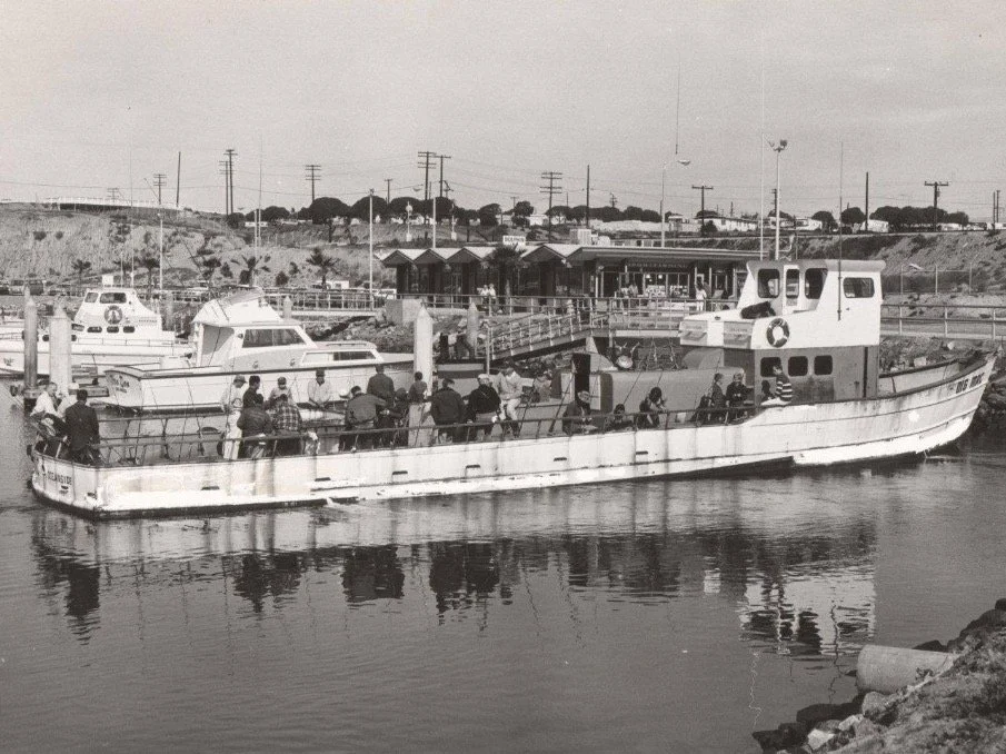 A black and white photo of a boat docked at a pier, carrying people boarding or disembarking, with buildings, power lines, and a hillside in the background.