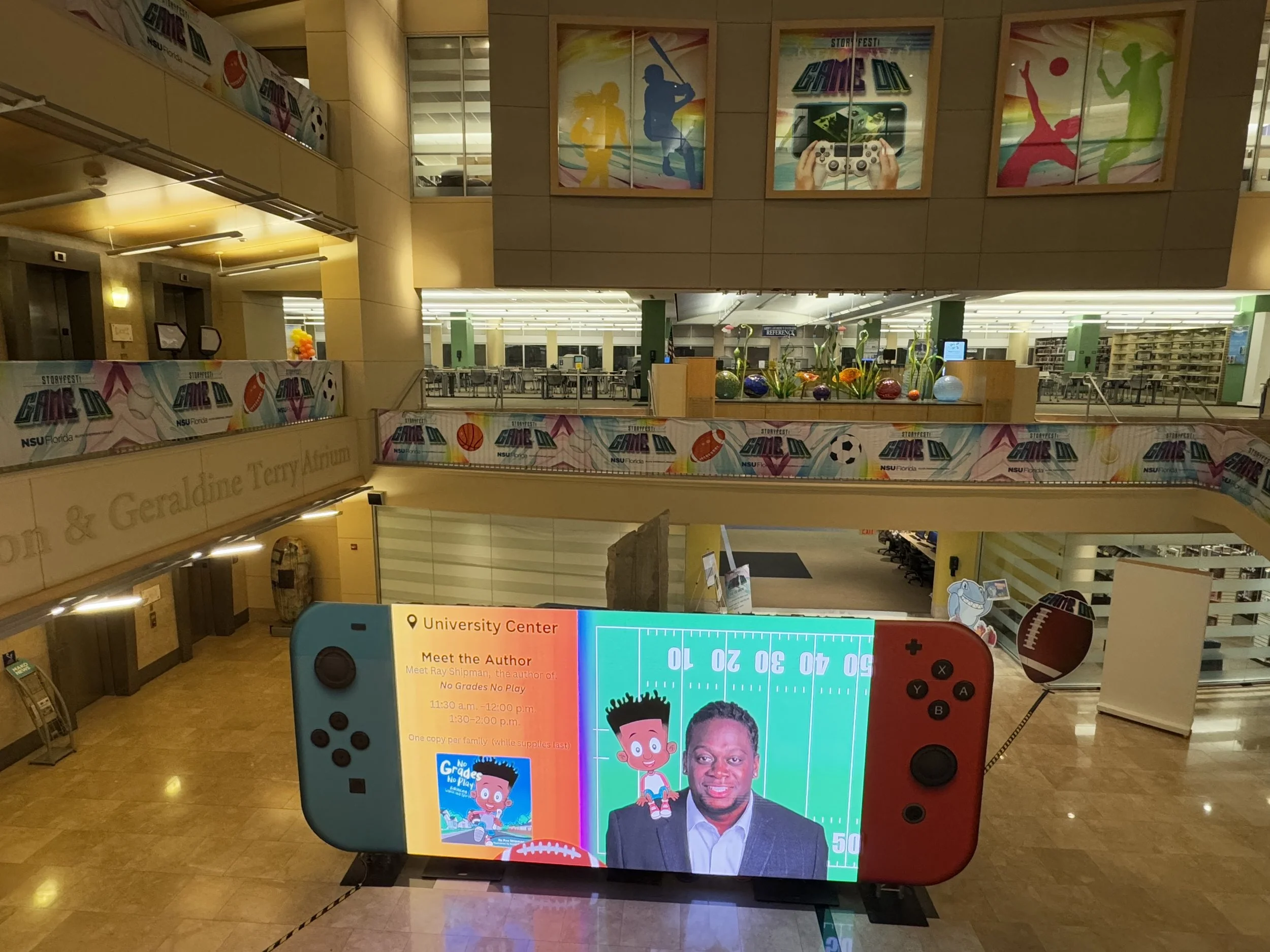Digital display shaped like a gaming console showing an author meet and greet event at the university center, with a photo of an author and a cartoon version of the same character, surrounded by posters and decorations celebrating gaming and sports.