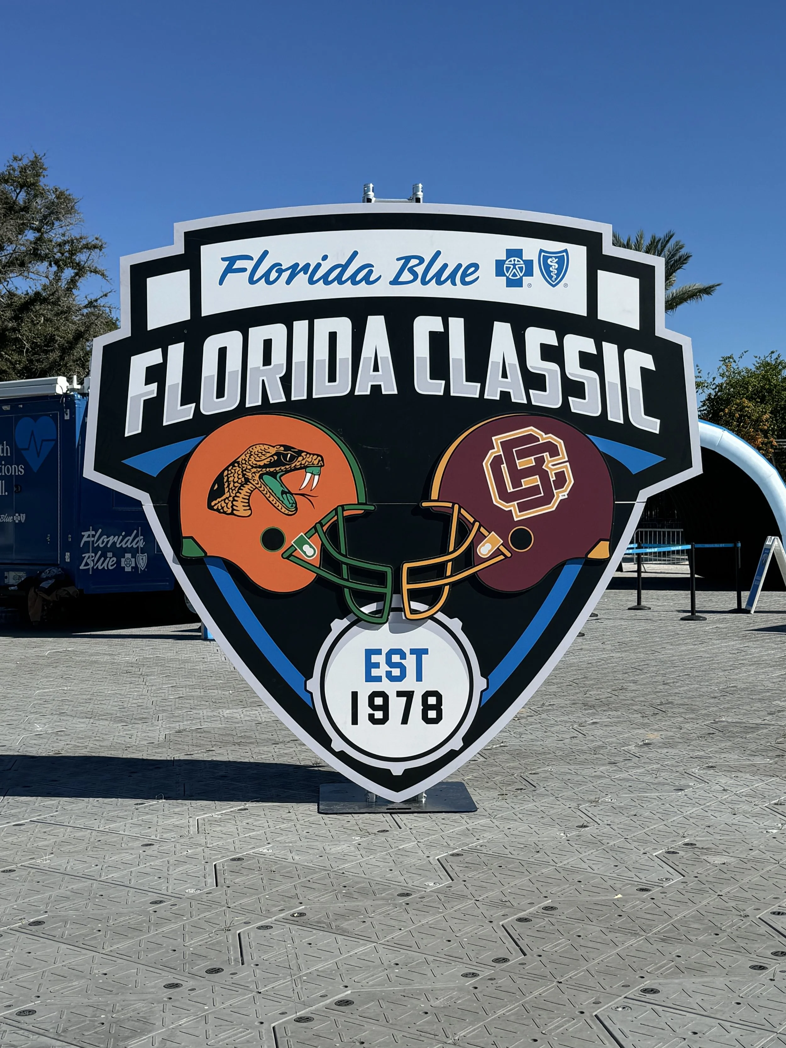 A large sign for the Florida Classic football game, featuring football helmets with a snake and a Gators logo, established in 1978, under the Florida Blue branding, set outdoors with a clear blue sky and palm trees in the background.