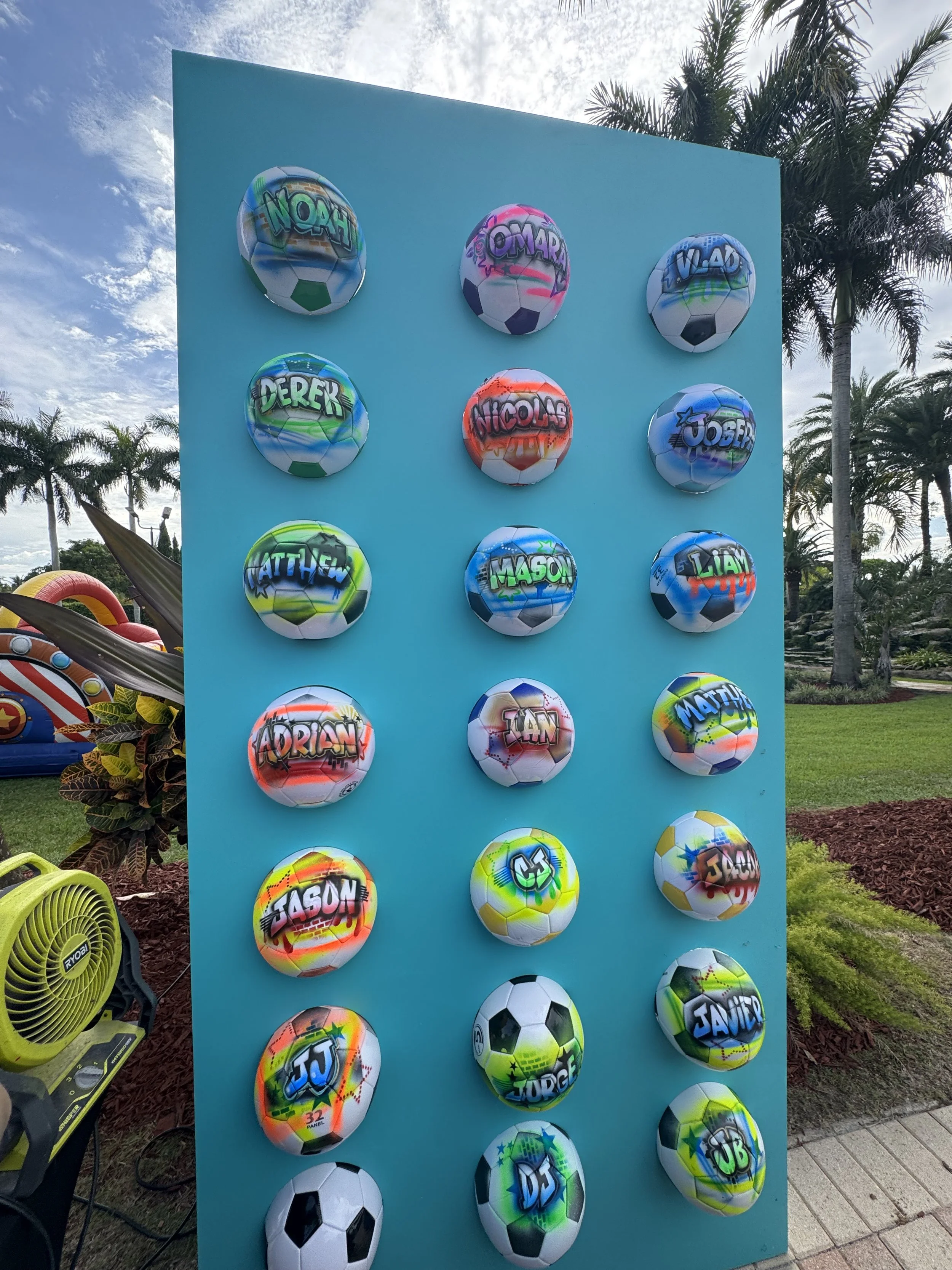 Colorful soccer balls with graffiti-style names and designs arranged on a blue wall outdoors, with palm trees in the background.