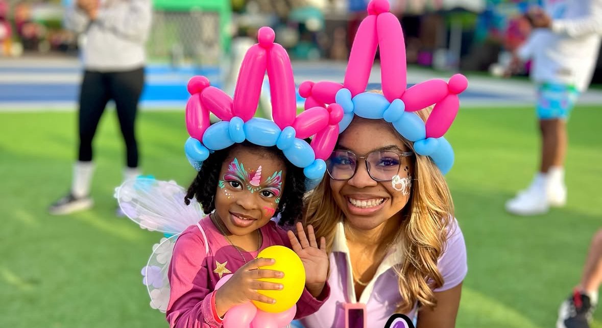 A young girl and a woman wearing balloon bunny ear headbands and smiling at the camera at an outdoor event, with colorful face paint on her face.