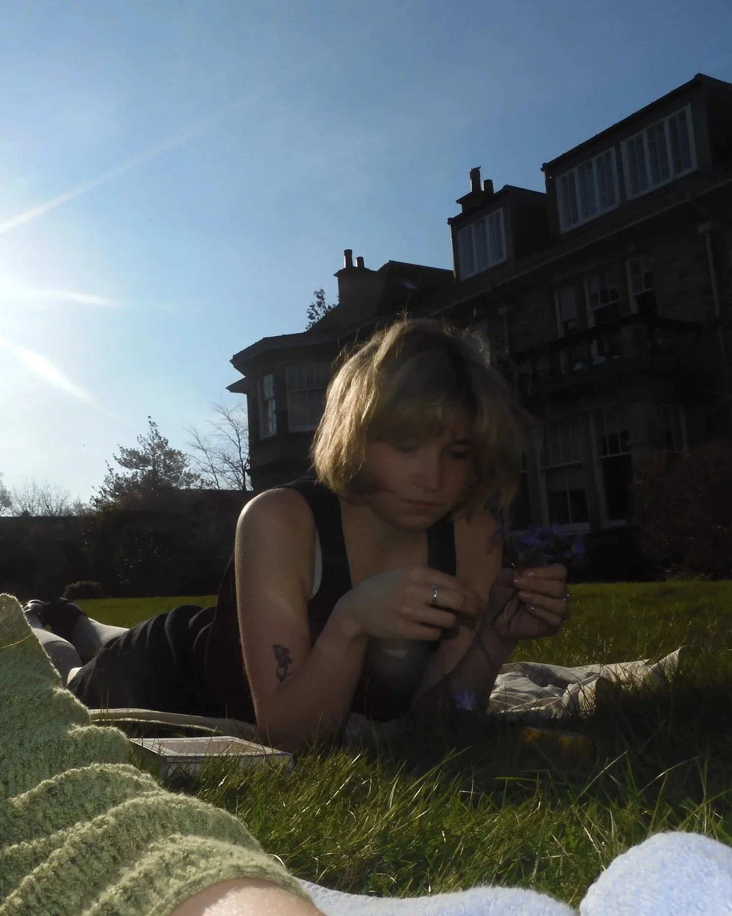 Ava Gomez laying on a grassy lawn, holding a flower, with a large house and a clear blue sky in the background.