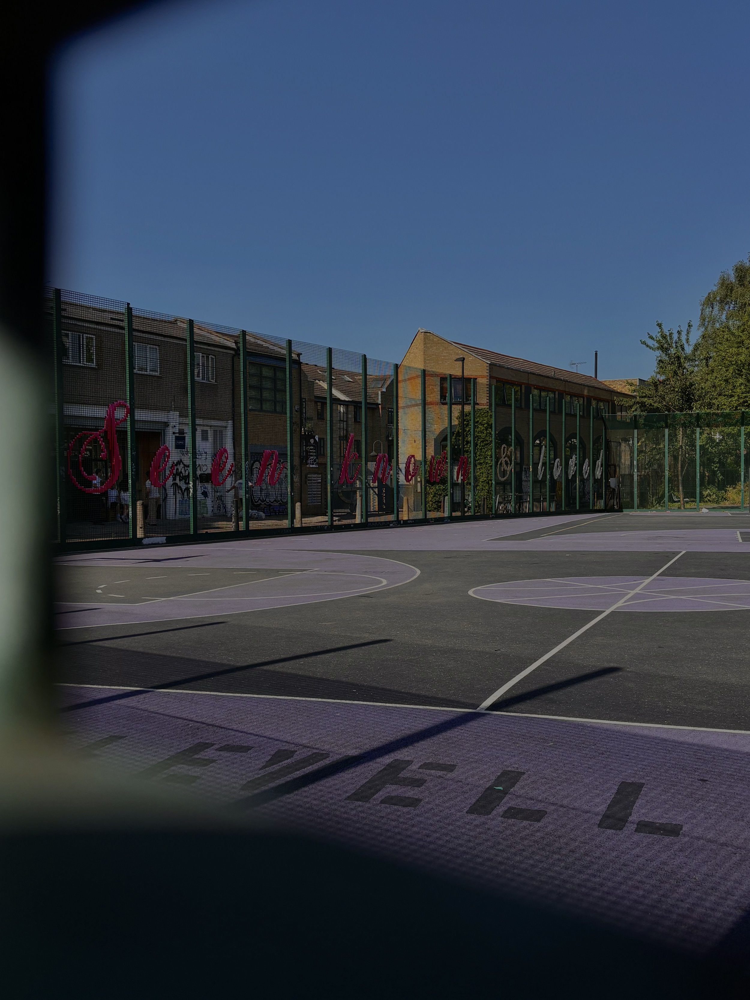 Empty basketball court with painted lines, surrounded by a fence with graffiti, residential buildings in the background, clear blue sky.
