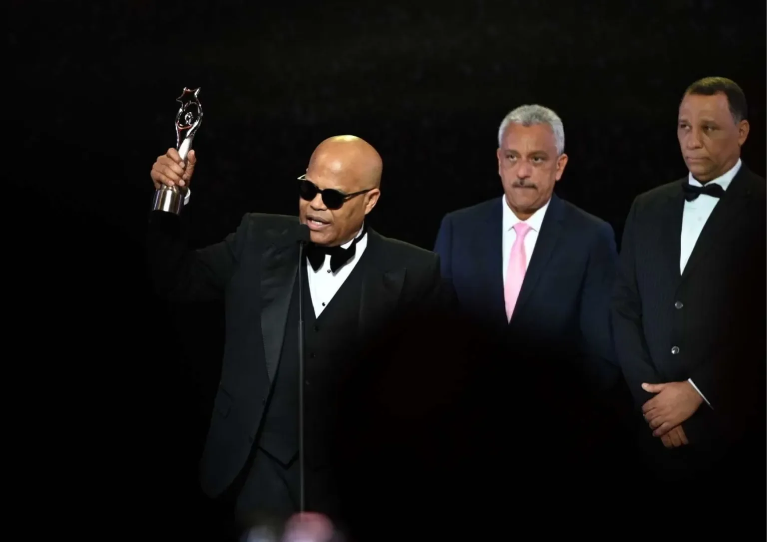 Man in a tuxedo with sunglasses is speaking at a podium, holding a trophy, flanked by two men in suits, during an awards ceremony.