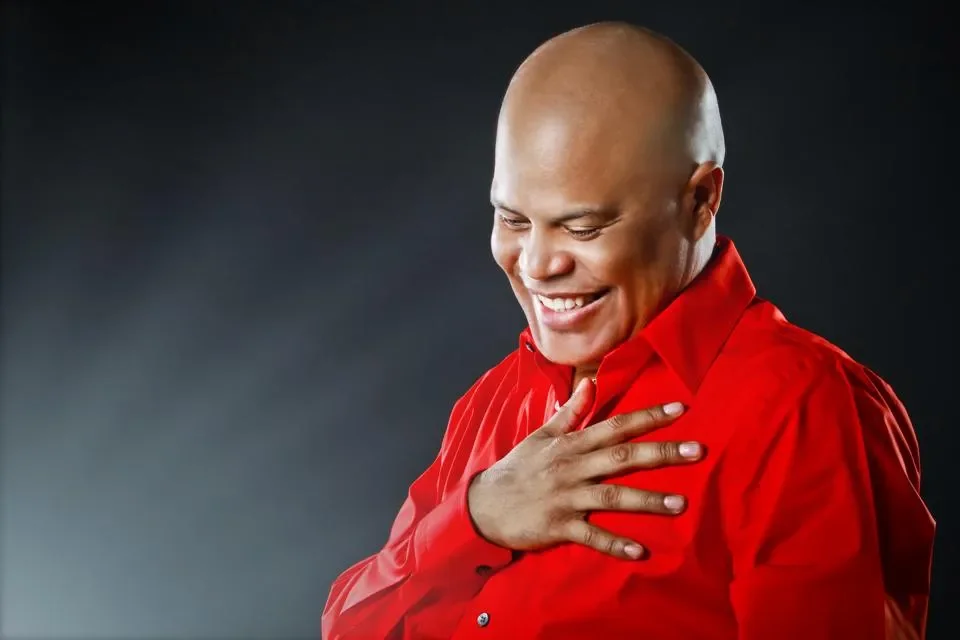 Rubby Perez smiling in a red shirt holding his chest with his right hand, standing against a dark background.