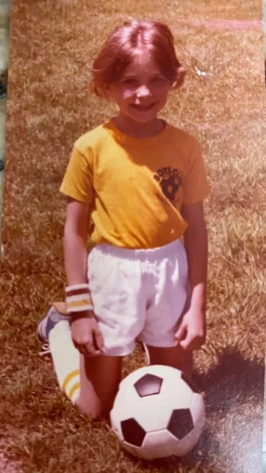 A young girl with red hair in a yellow sports shirt and white shorts is standing on a grassy field. She is smiling and has a soccer ball resting on her foot.