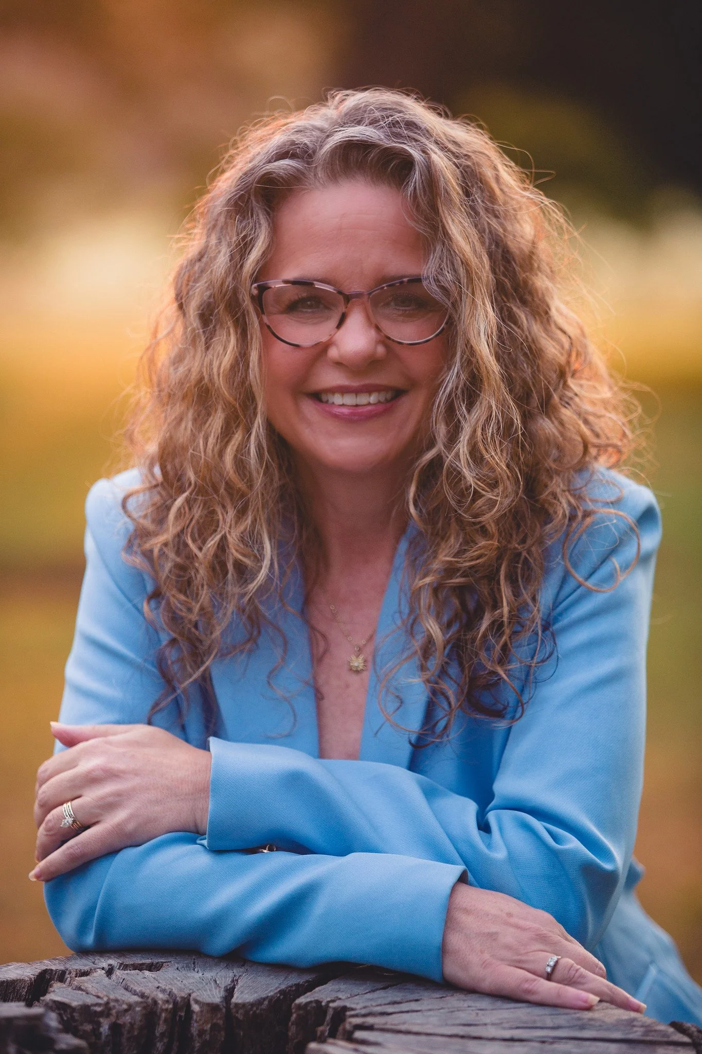 Woman with curly hair wearing glasses and a blue blazer, smiling outdoors with a blurred natural background.