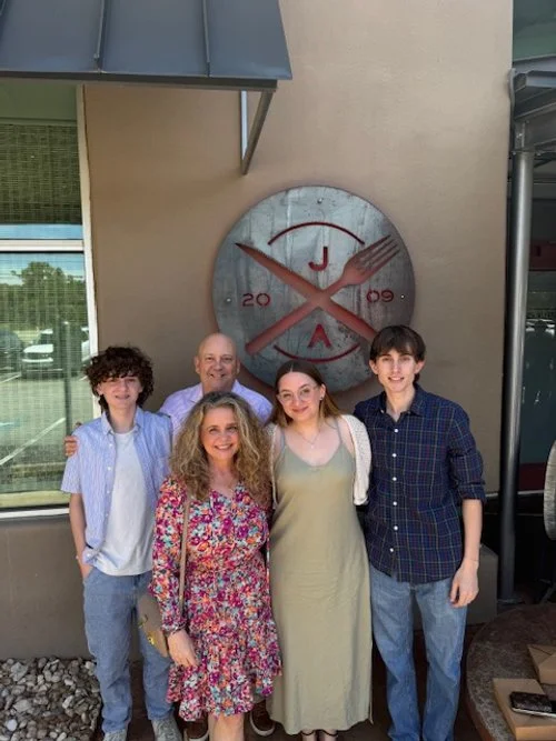 A group of six people standing in front of a large decorative wall clock with a fork and knife design, outdoors near a building.
