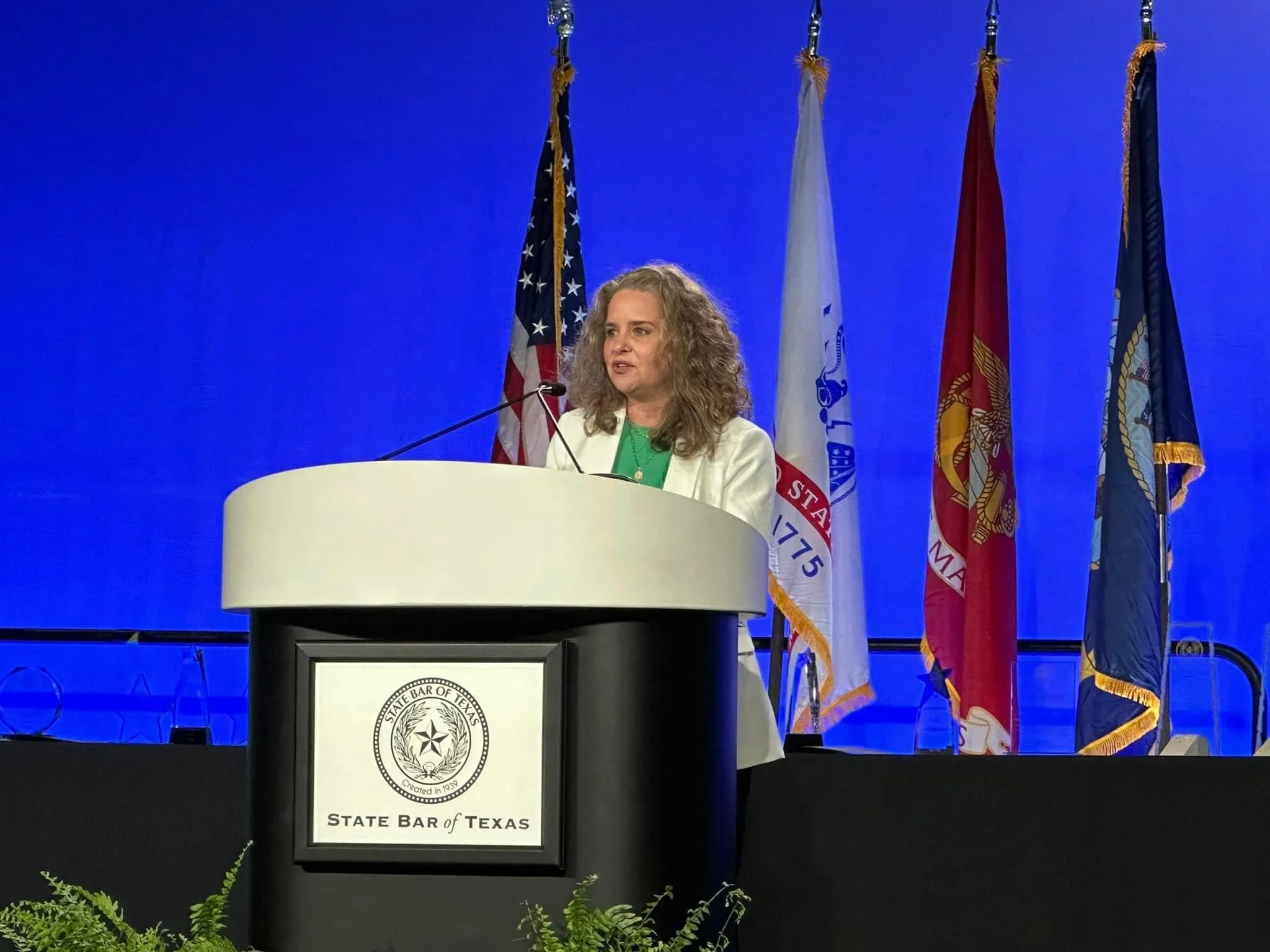 A woman with curly hair standing at a podium labeled "State Bar of Texas," speaking at a conference. Behind her are four flags and a blue background.