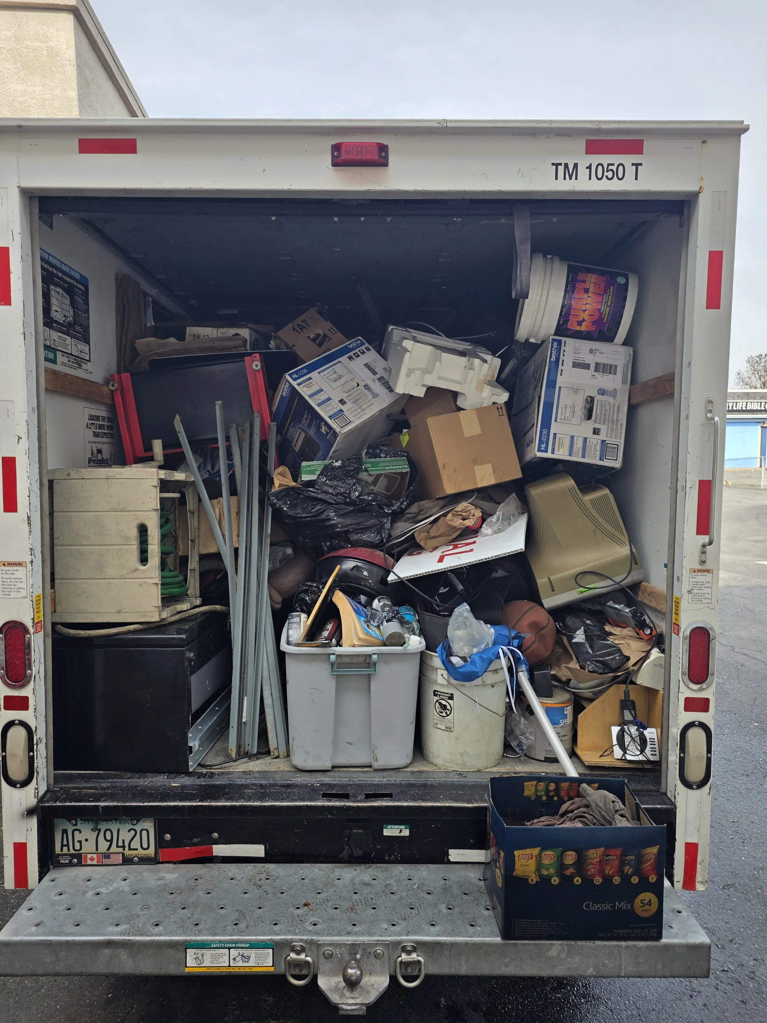 The back of a box truck filled with miscellaneous items including boxes, a cooler, household items, and trash.