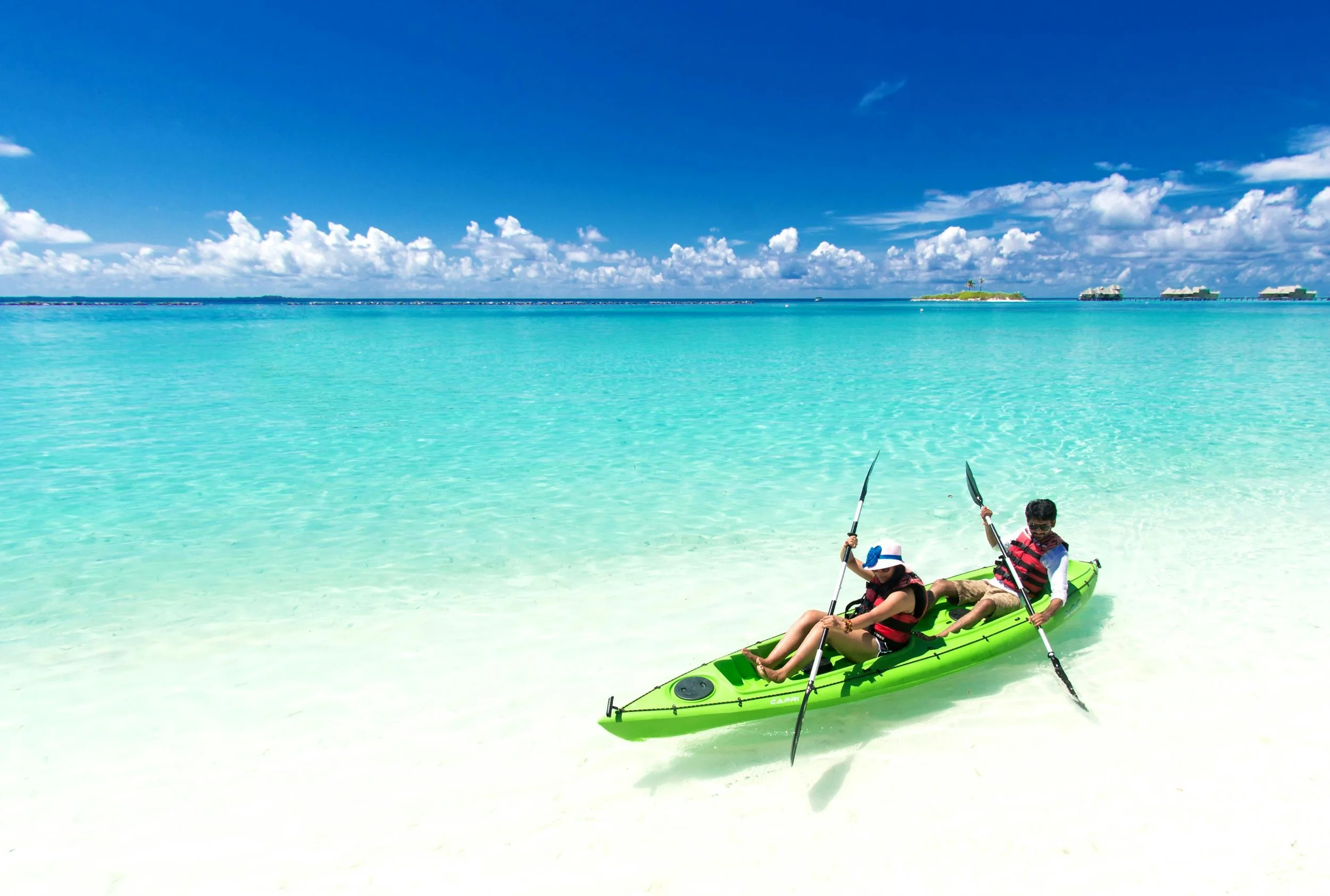 Two people in a green kayak on a white sandy beach with turquoise water under a blue sky with clouds.