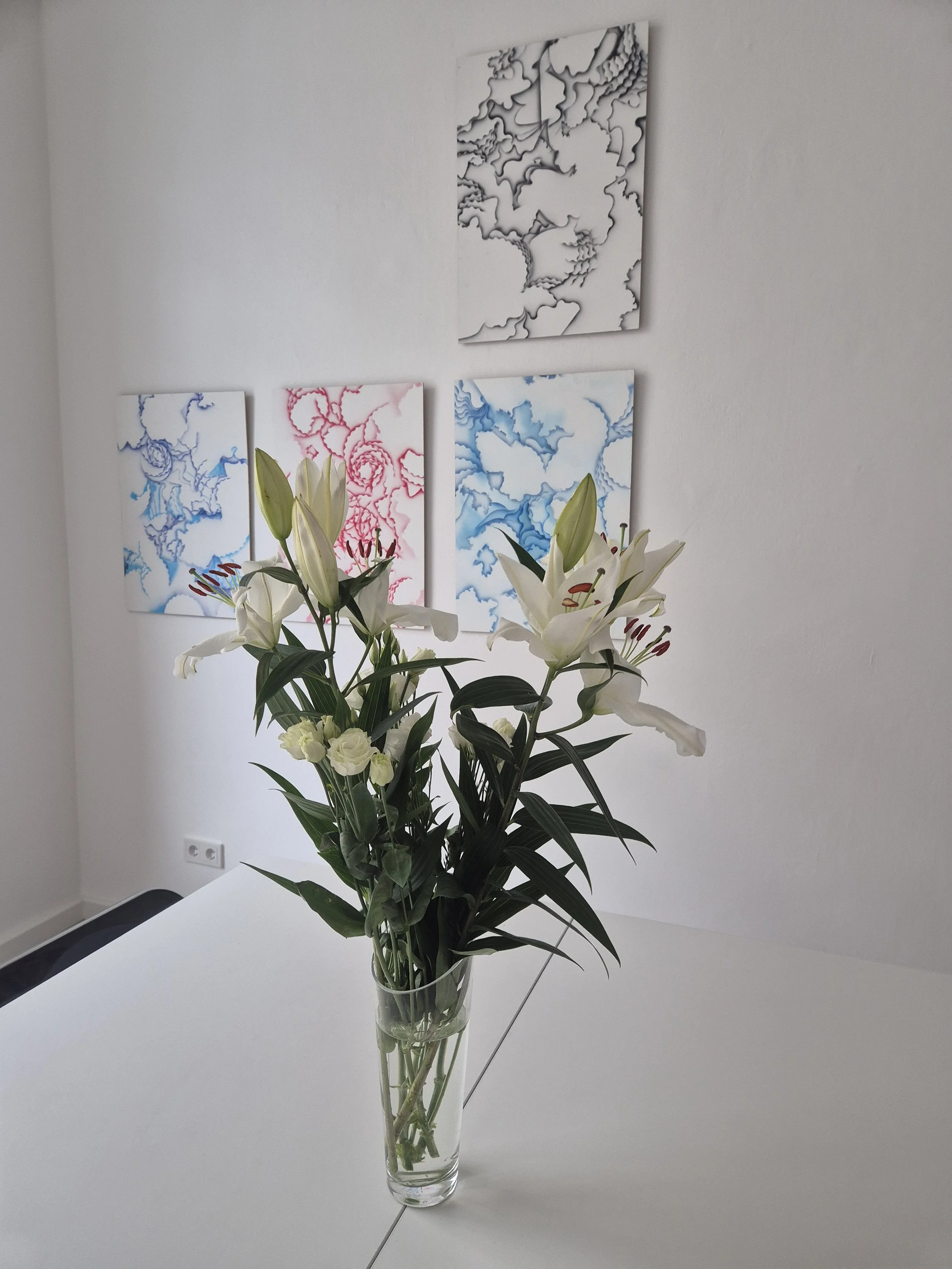 Blumenstrauß mit weißen Lilien und kleinen weißen Rosen in einer Glasvase auf einem weißen Tisch, im Hintergrund Wand mit abstrakten Kunstwerken in Blau, Pink und Schwarz.