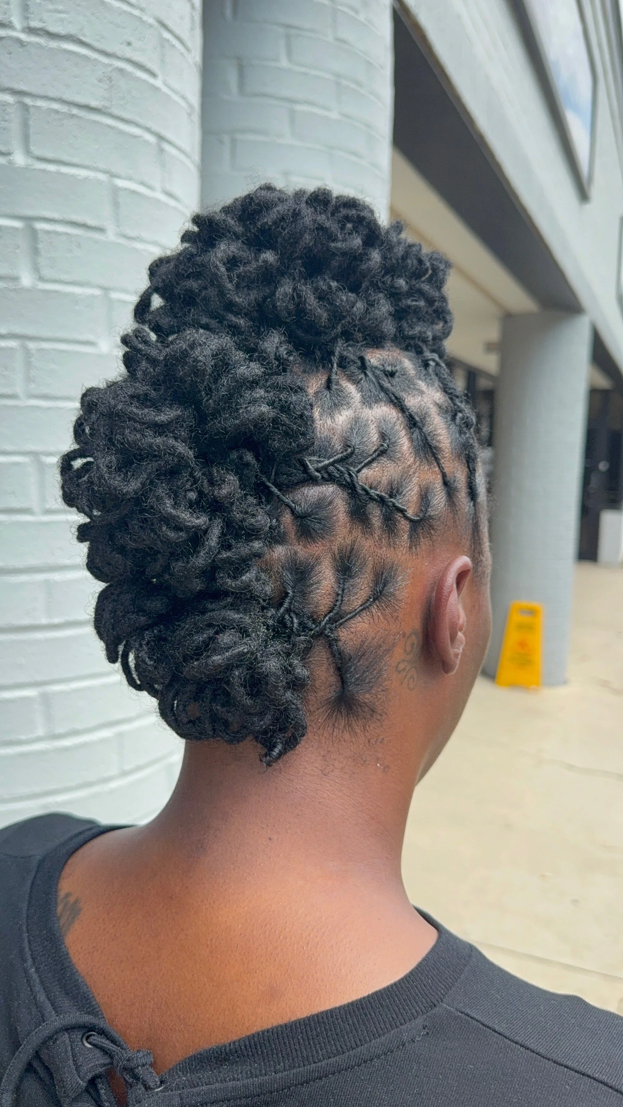 Back of a person's head with black, textured dreadlocks styled into a high bun, with shaved sides and intricate patterns shaved into the scalp.